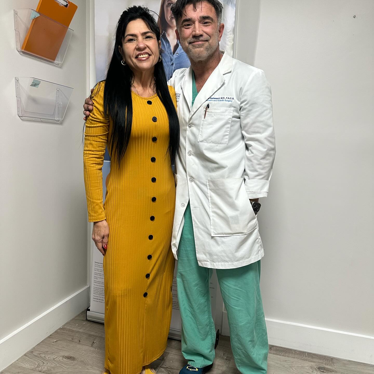 Smiling gastric sleeve patient in a yellow dress standing with Dr. Reza Keshavarzi in the office at her one-year milestone