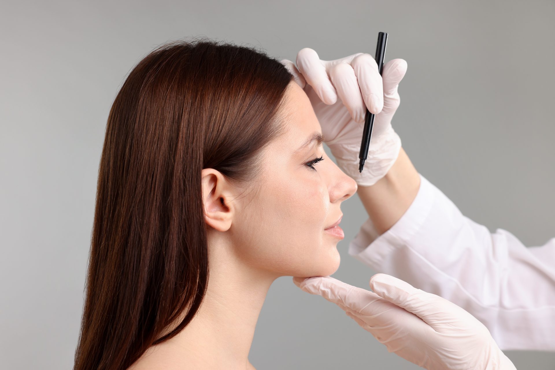A gloved provider marks a woman's nose in profile during a rhinoplasty consultation, shown against a plain gray studio backgr