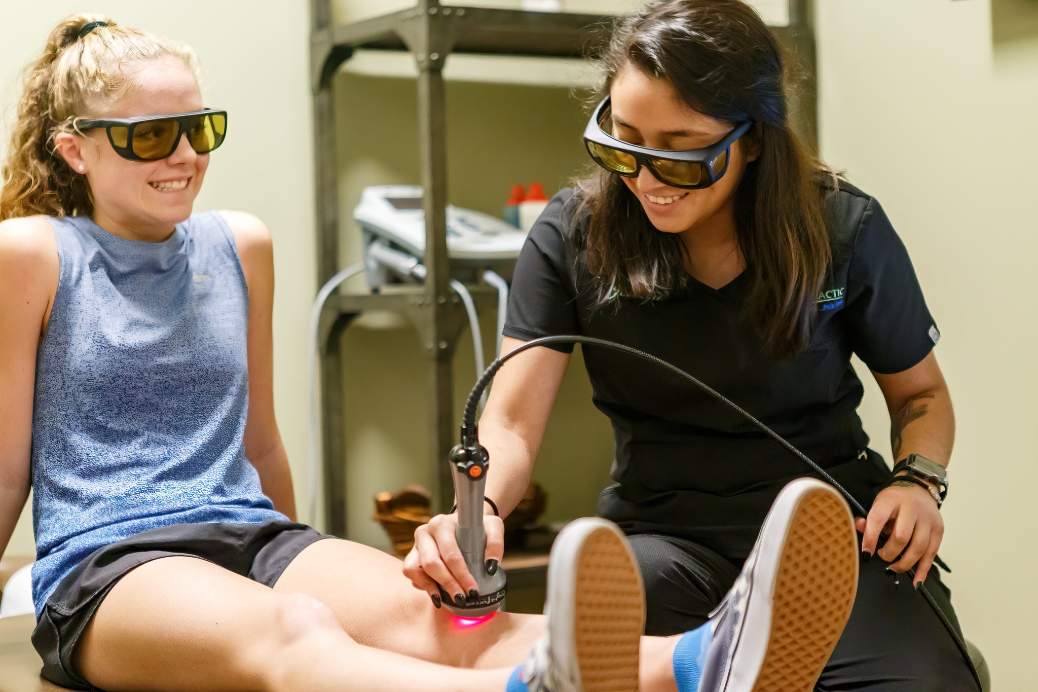 A clinician in dark scrubs uses a cold laser device on a smiling patient's knee while both wear protective eyewear