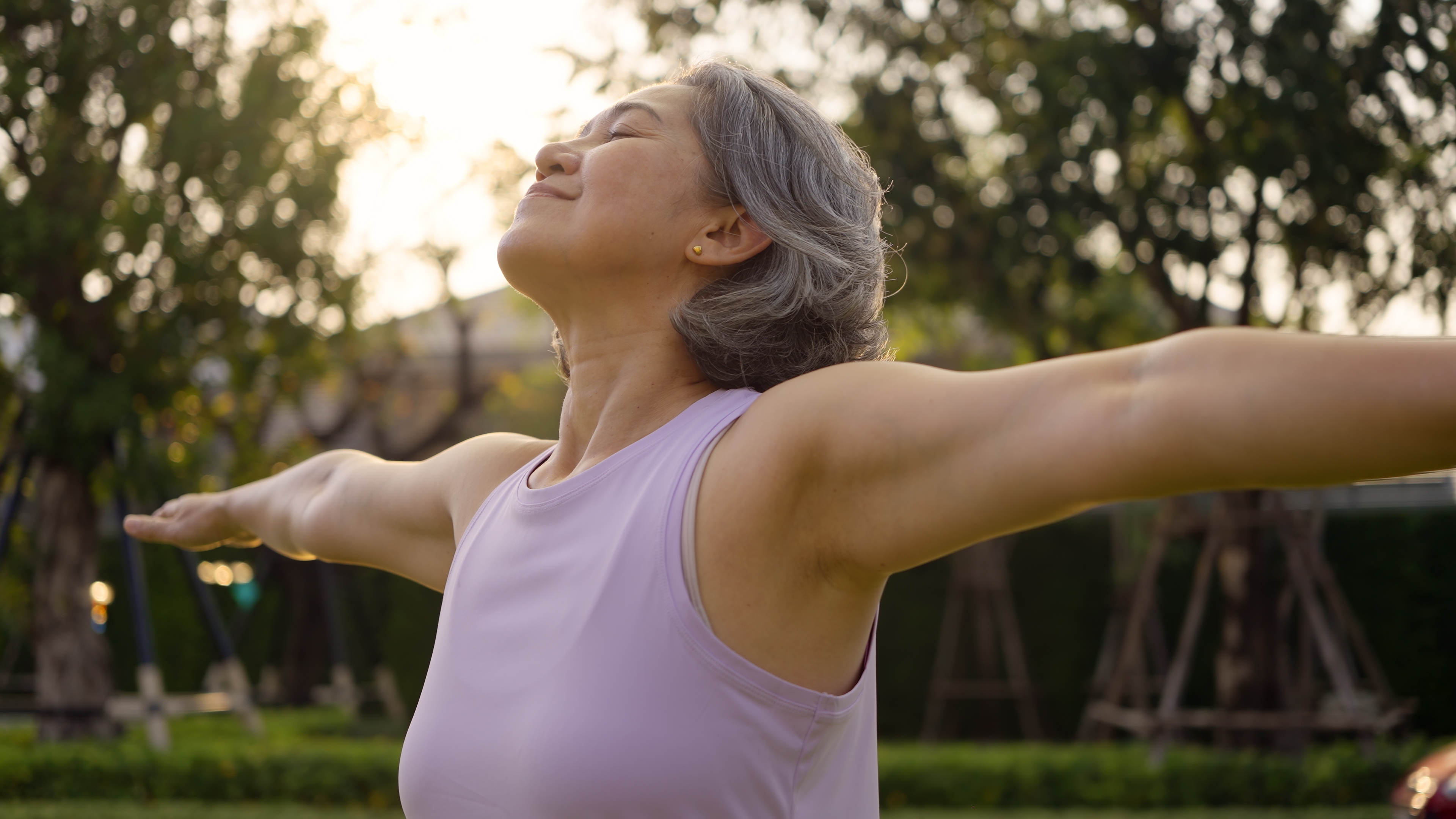 Older woman with arms spread wide outdoors among trees, eyes closed, enjoying good health