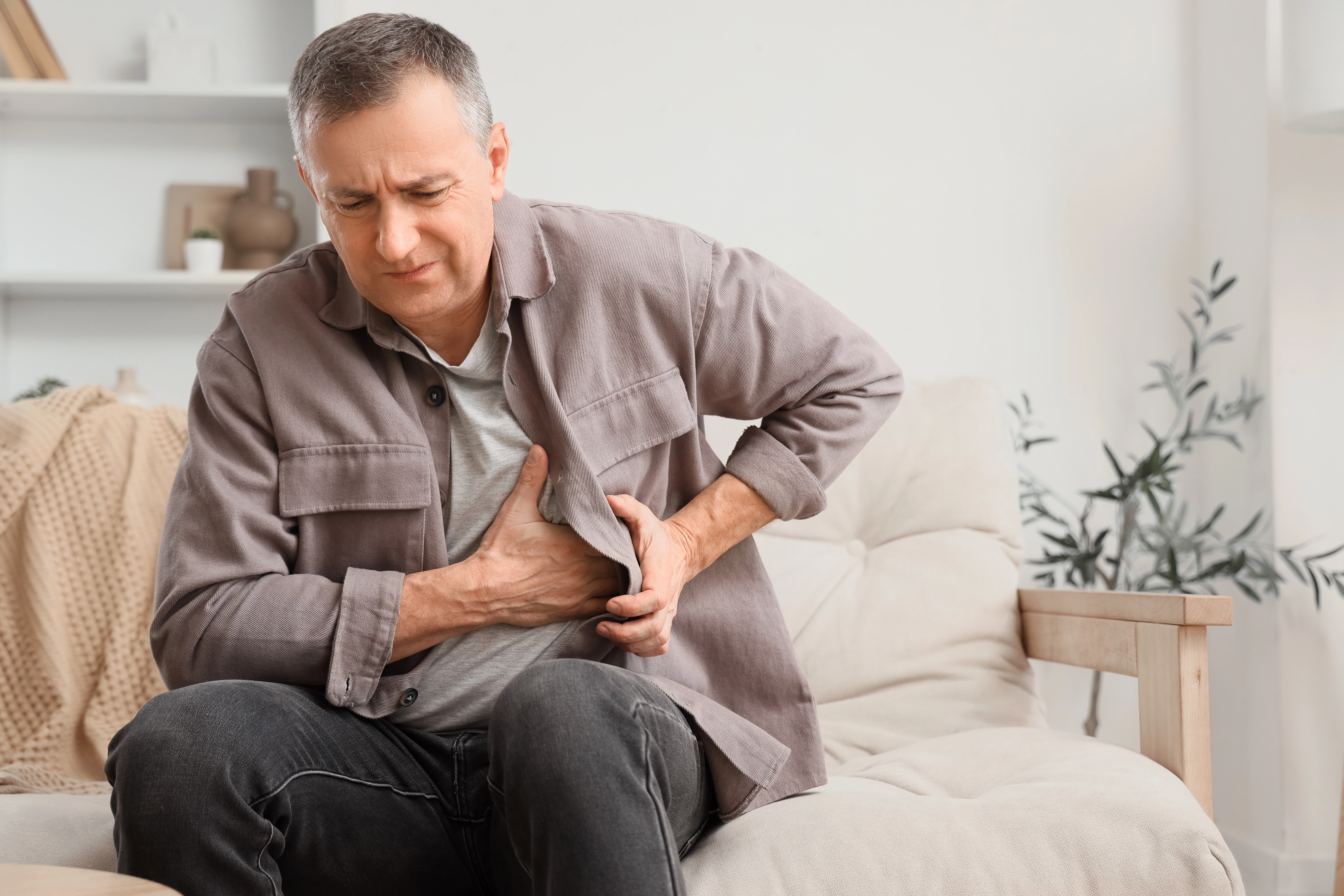 A middle-aged man sits on a sofa clutching his chest in discomfort, illustrating a possible sign of a heart condition needing