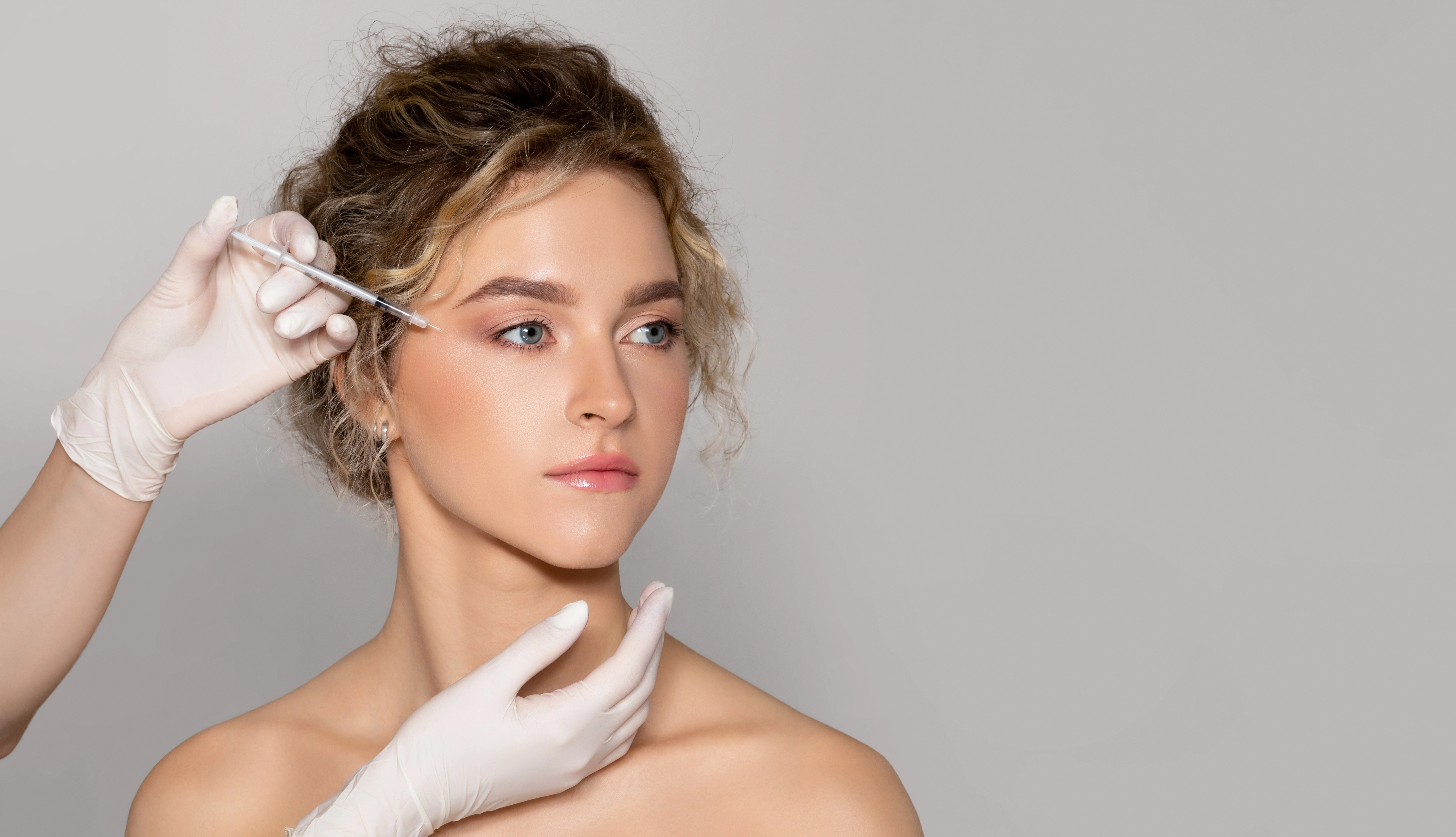 A gloved provider administers a facial injection near the eye of a young woman against a neutral studio background