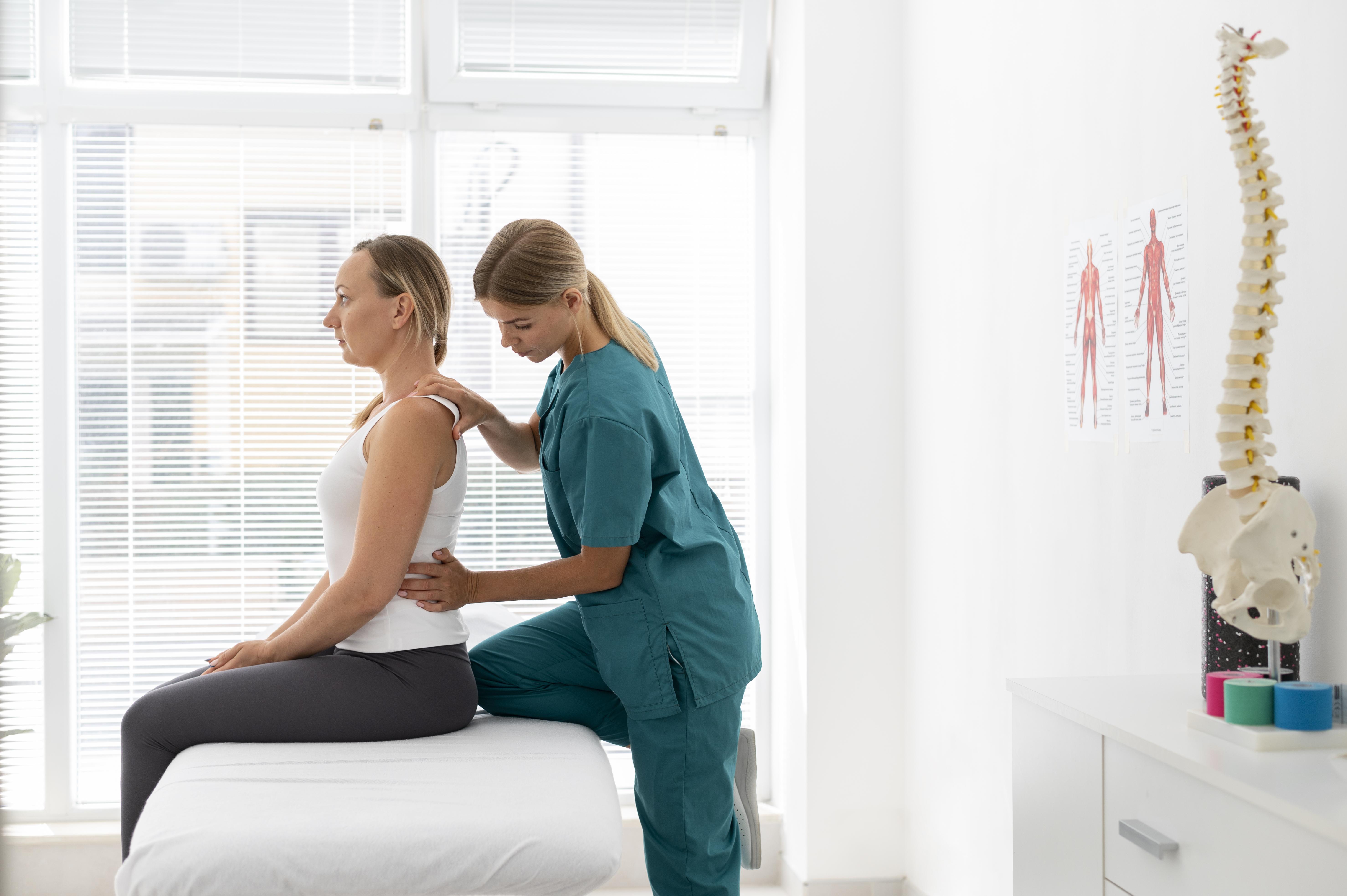 A chiropractor in teal scrubs adjusts a seated female patient's back in a bright clinical treatment room with a spine model