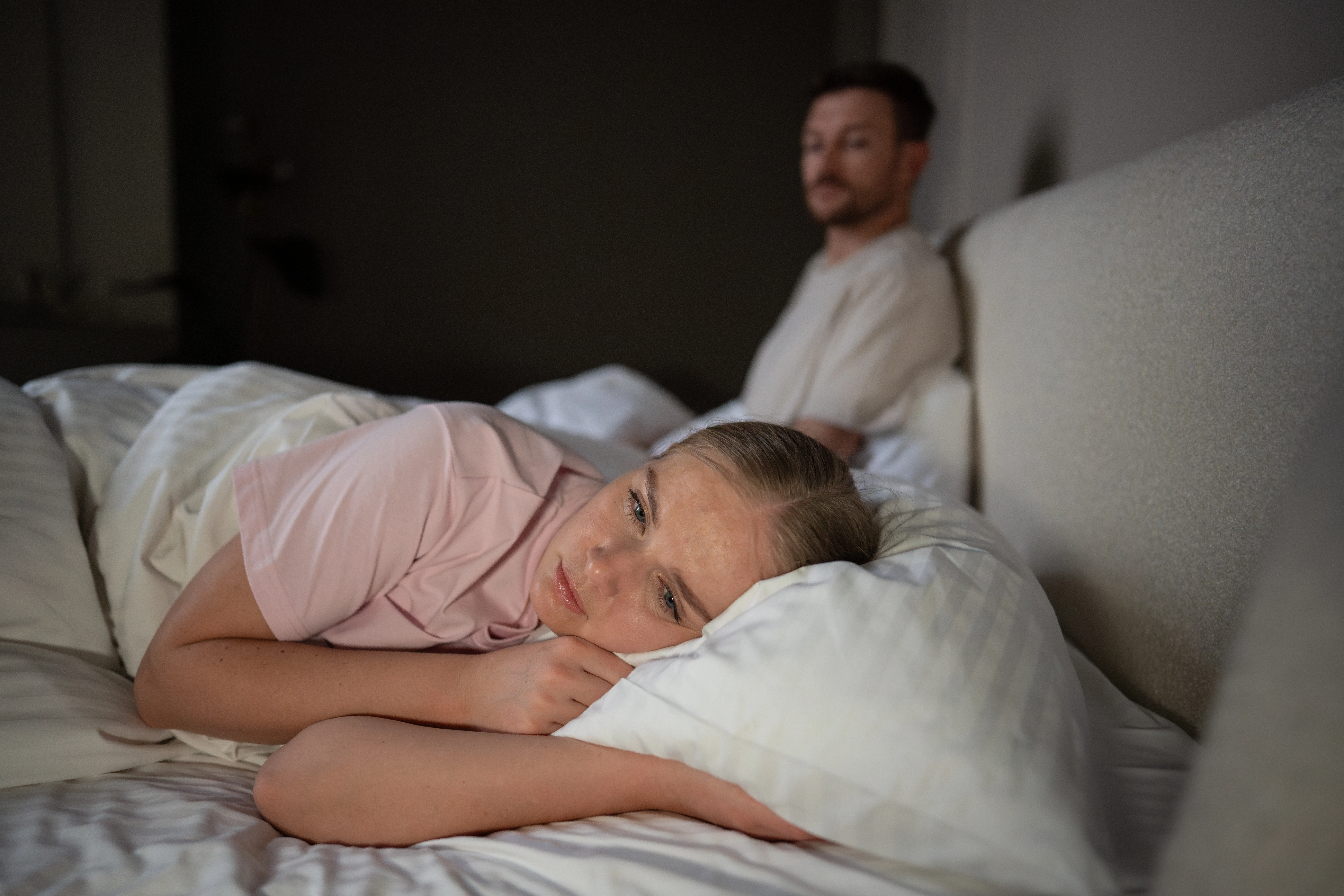 A woman lies awake looking distressed in bed while a man sits behind her, illustrating relationship distance and low libido i