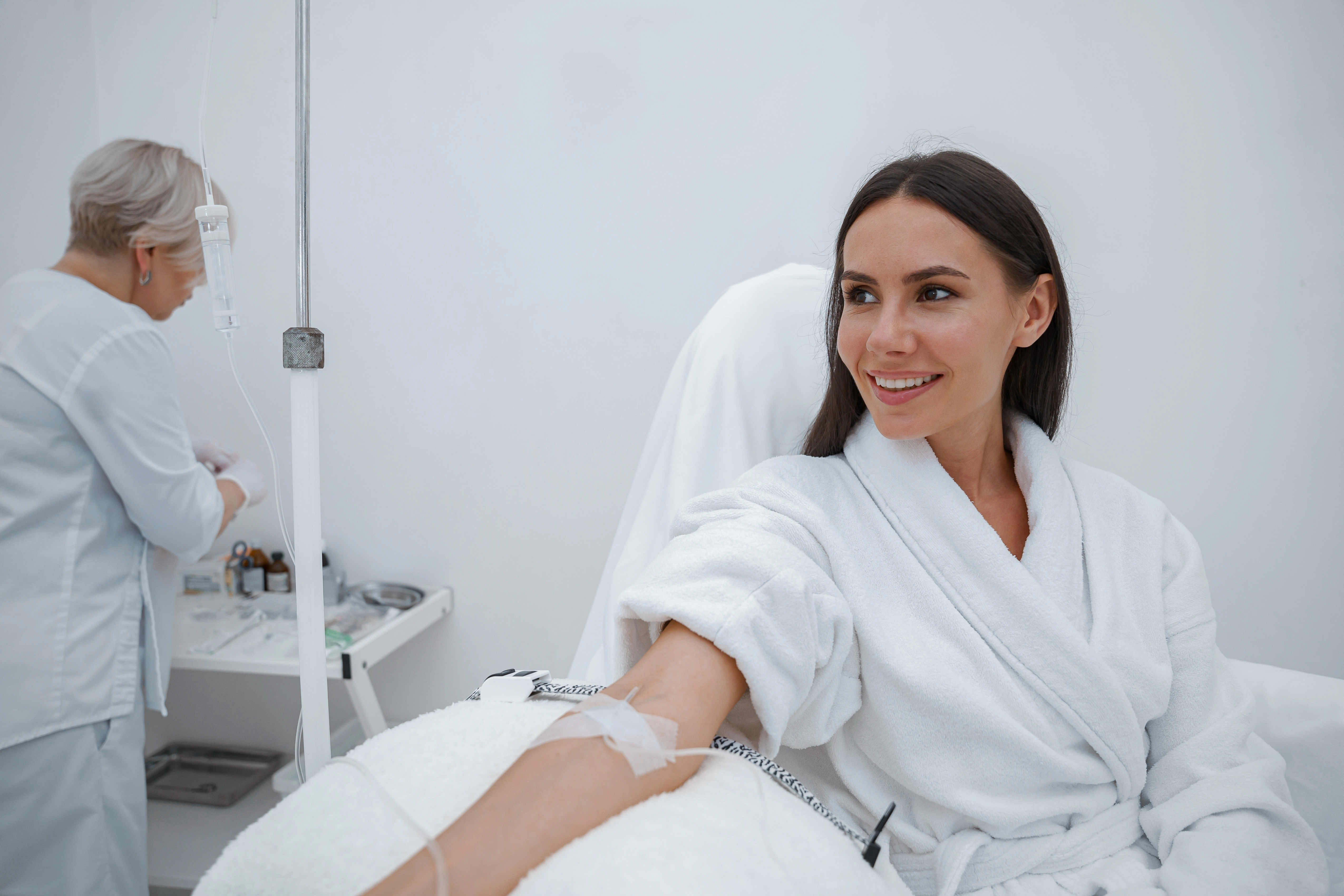 A smiling woman in a white robe receives an IV infusion while a provider prepares supplies in the background.