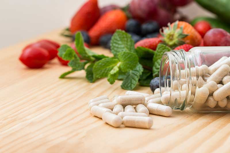 Open jar of white supplement capsules on a wooden table beside fresh strawberries, blueberries and mint