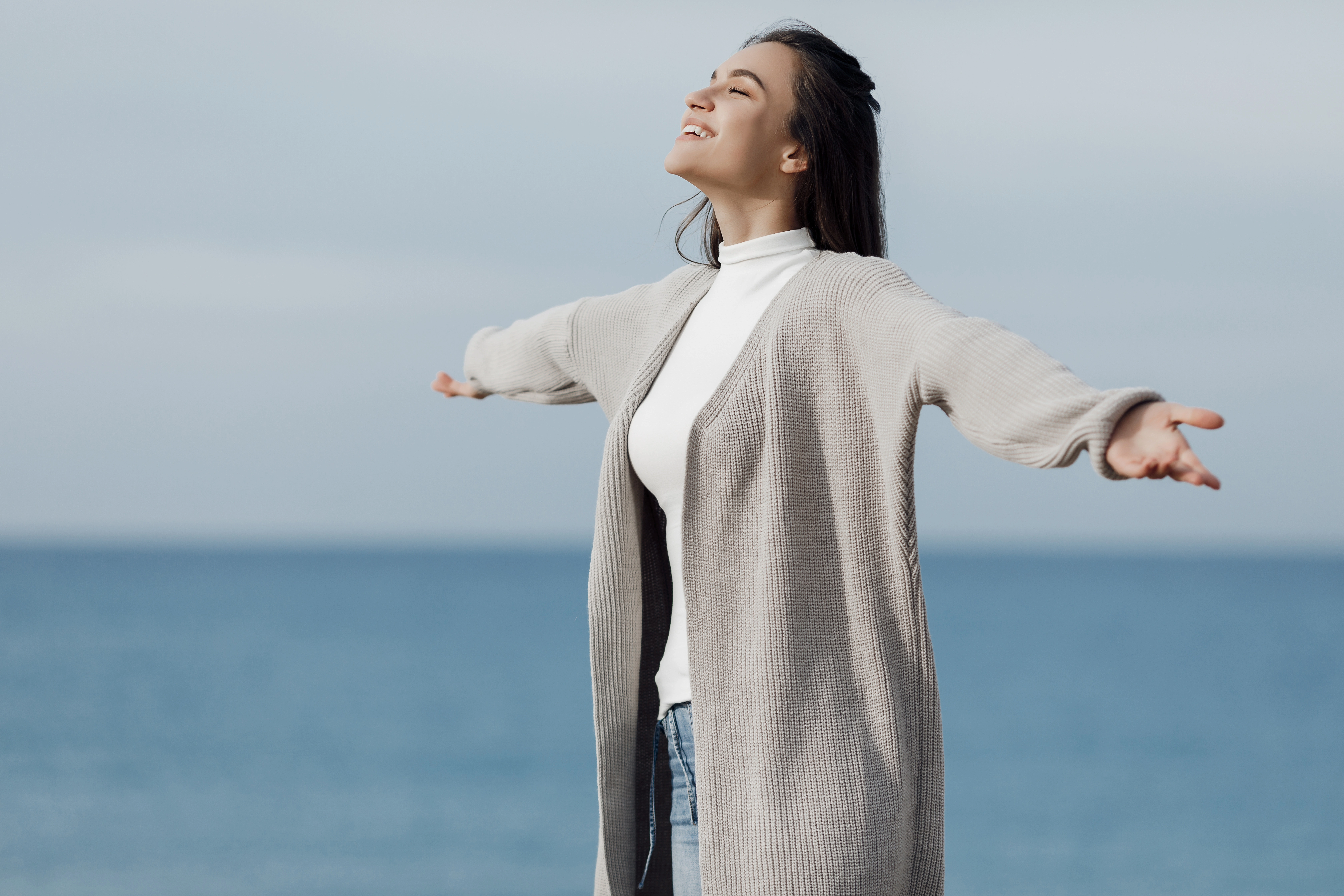 Woman in a light cardigan breathing deeply with arms open beside the sea