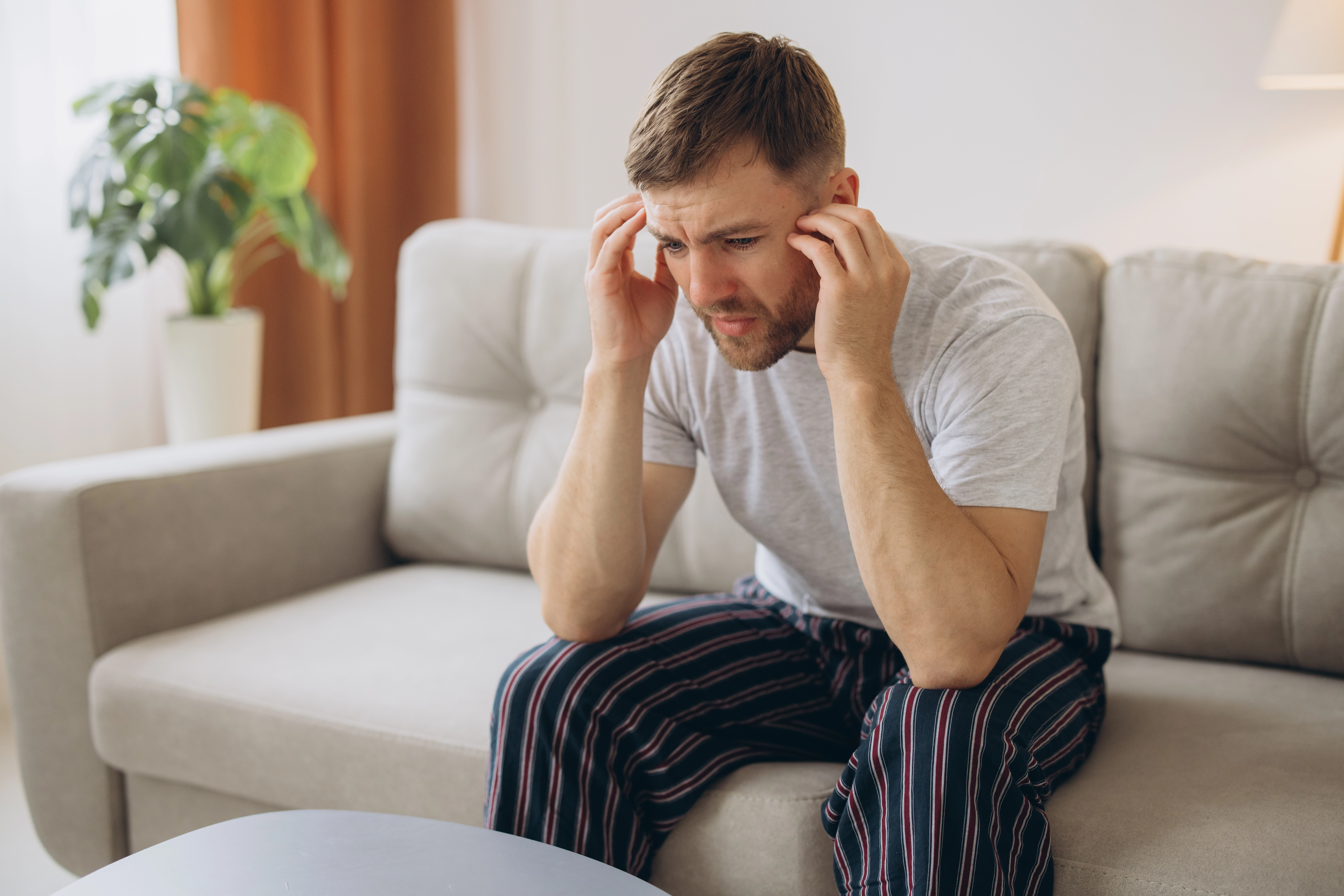A distressed man sits on a couch at home holding his head, illustrating stress or anxiety symptoms related to PTSD