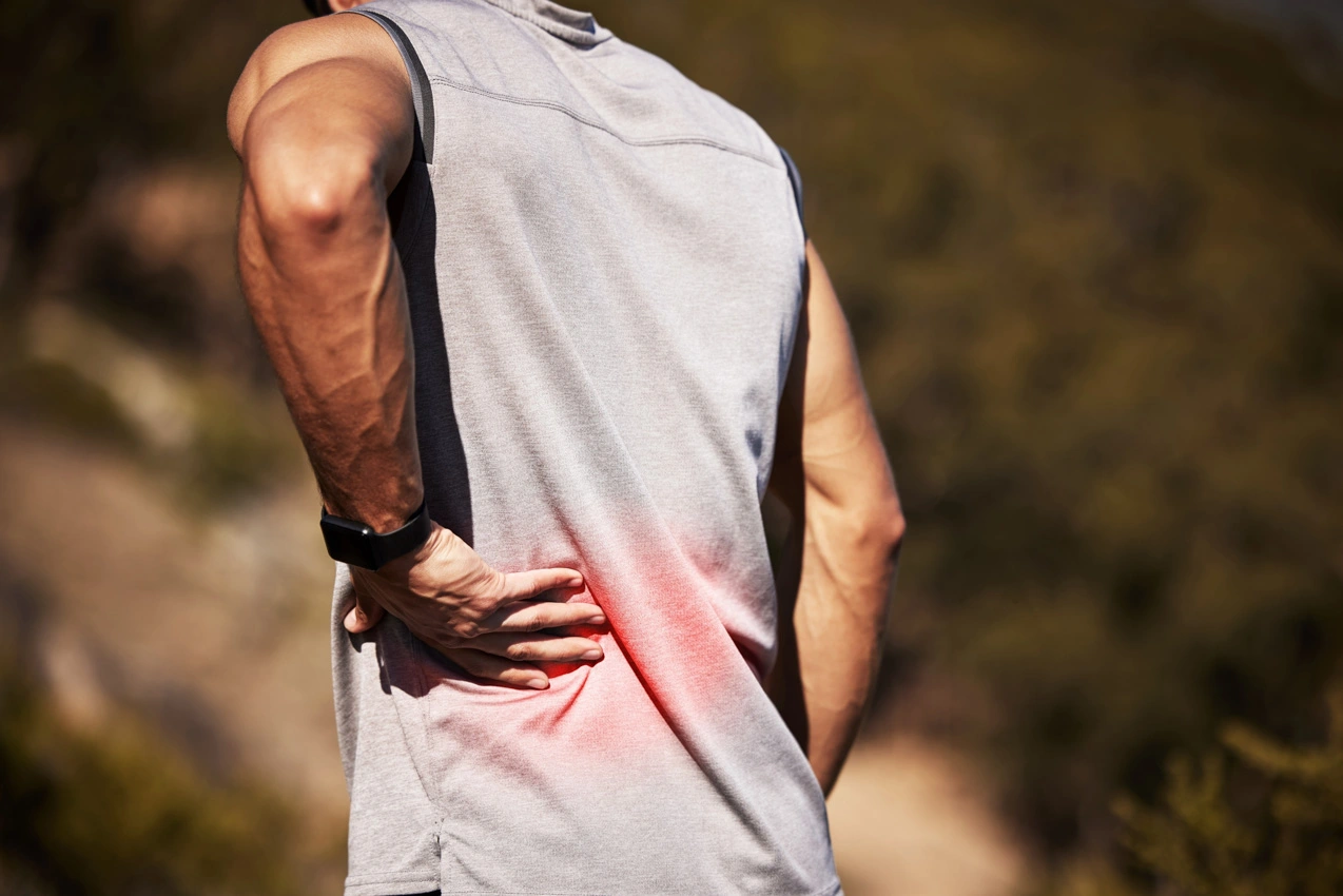 A man outdoors grips his lower back in pain, highlighted with a red graphic overlay indicating discomfort.