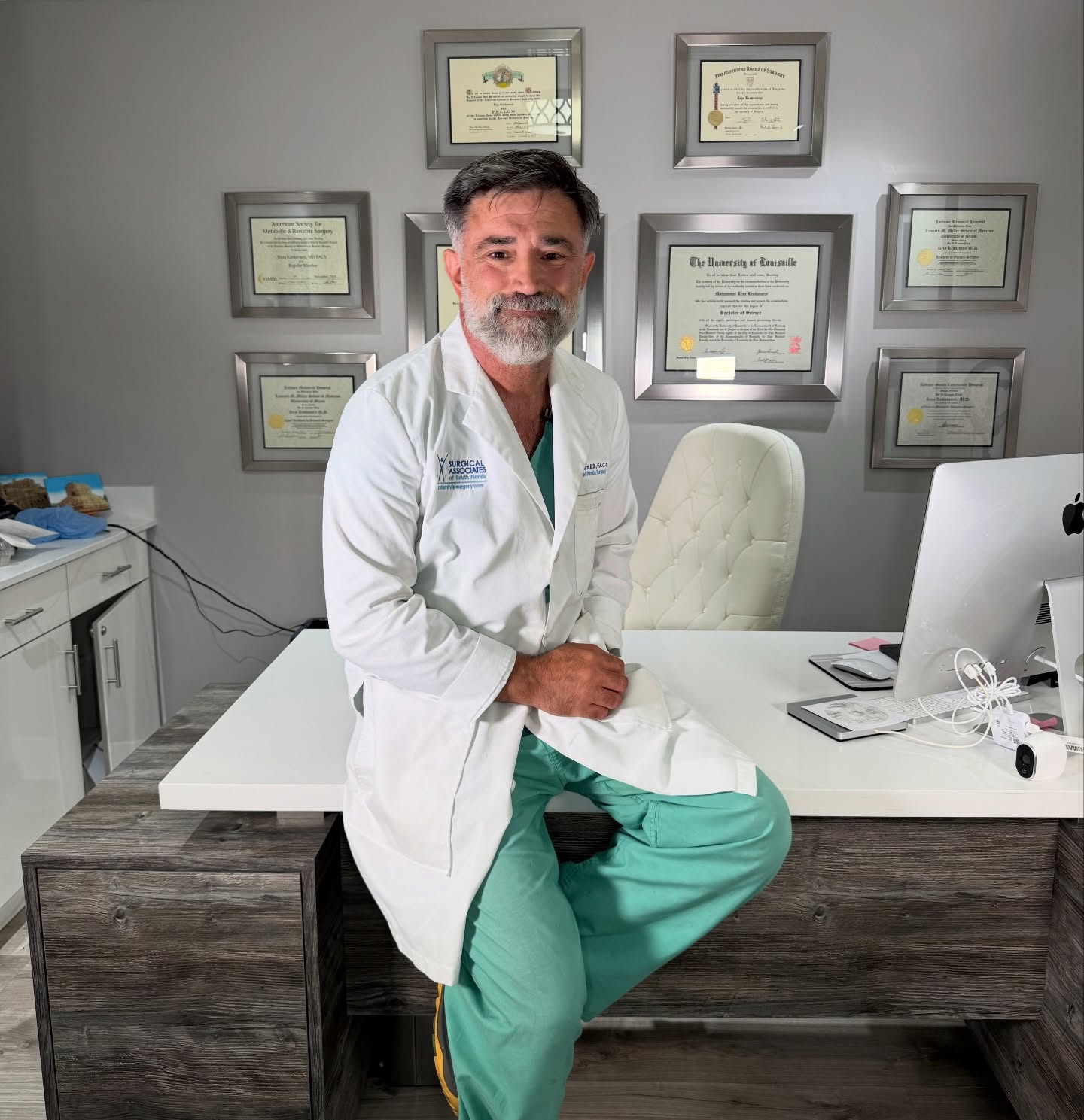 Dr. Reza Keshavarzi in a white coat seated on his desk in front of his framed diplomas