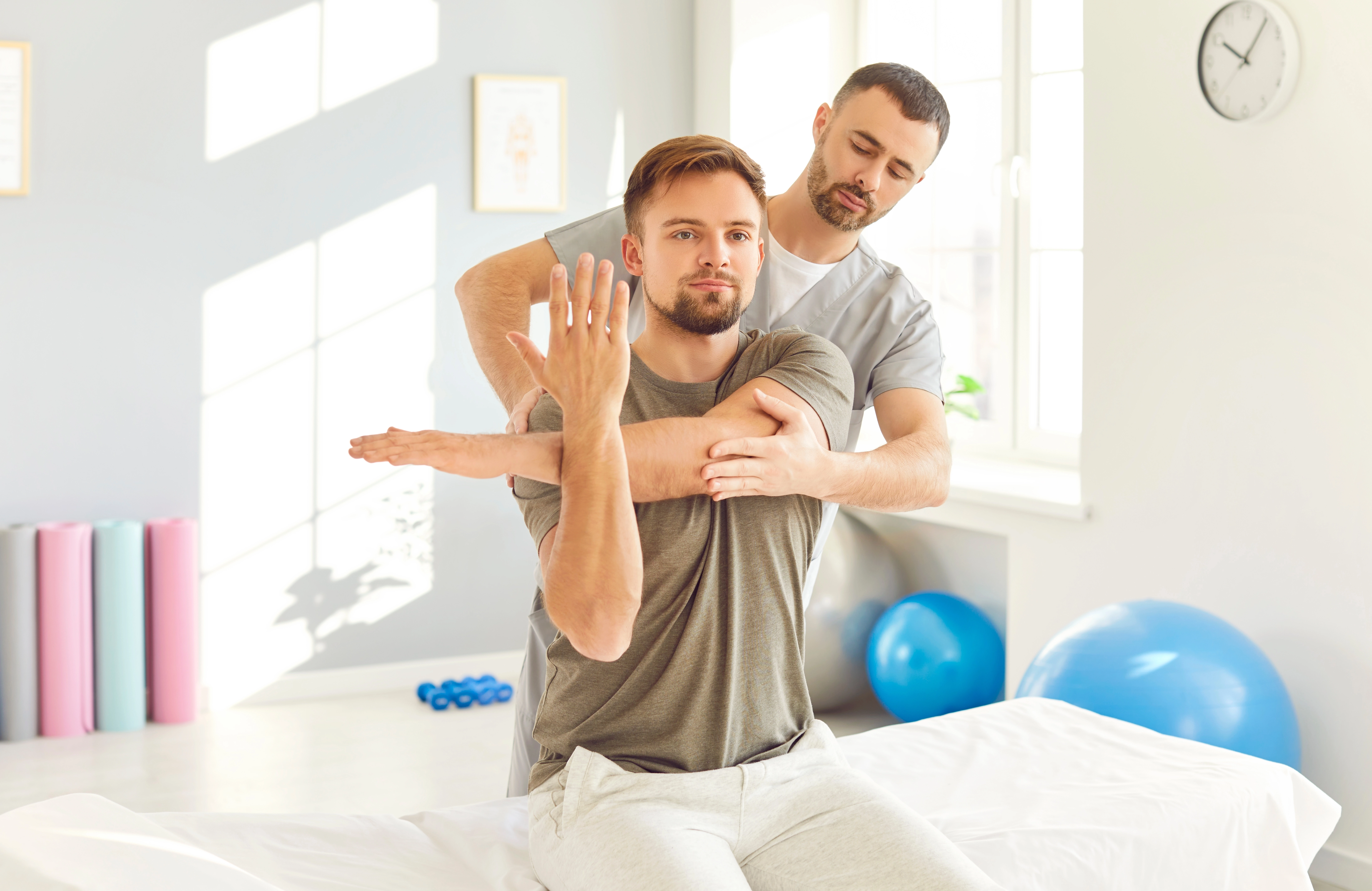 A chiropractor assists a male patient with an arm and shoulder stretch during a chiropractic evaluation in a bright clinical setting