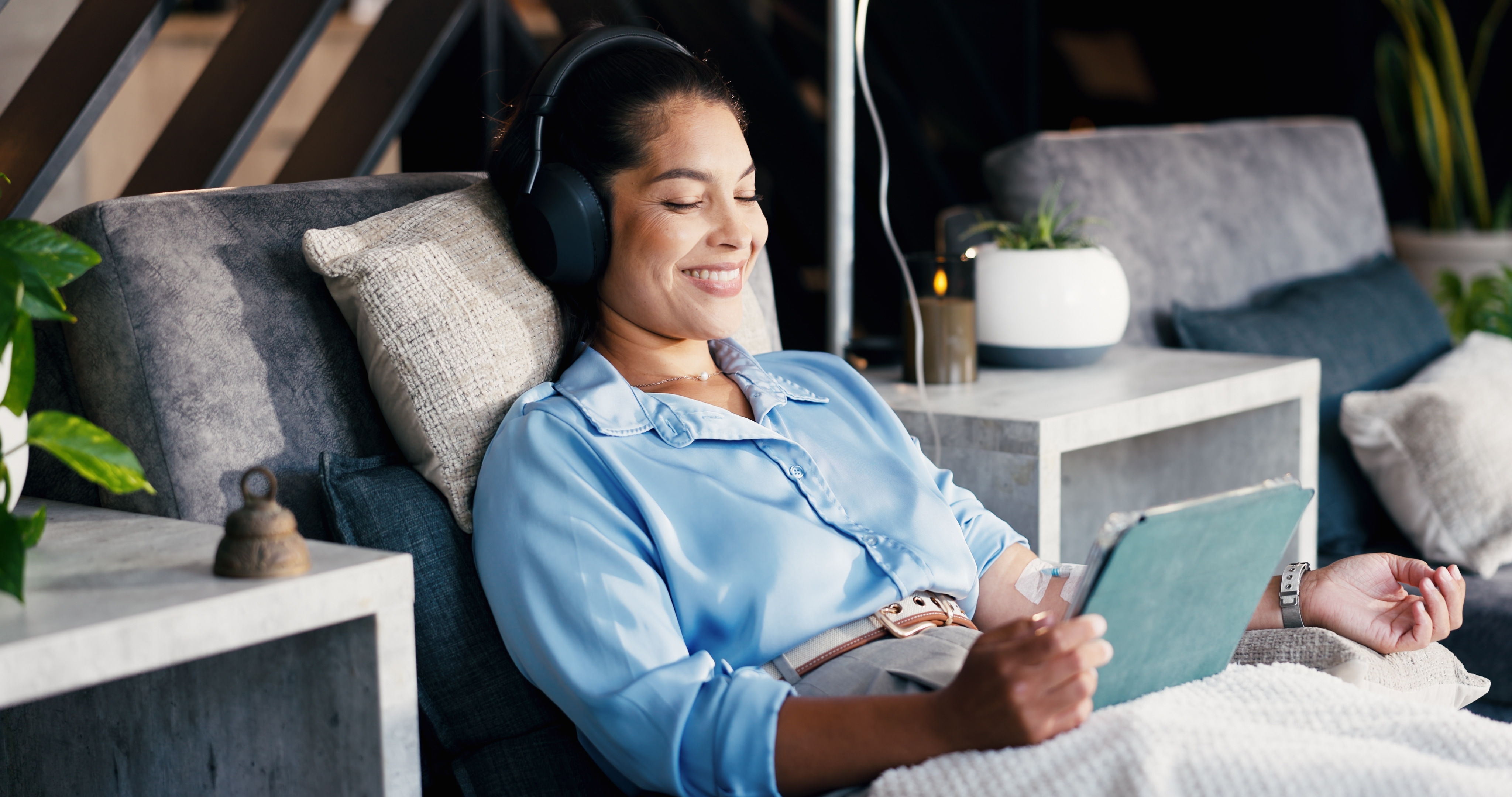 Smiling woman relaxing on a sofa with headphones and a tablet during an IV infusion