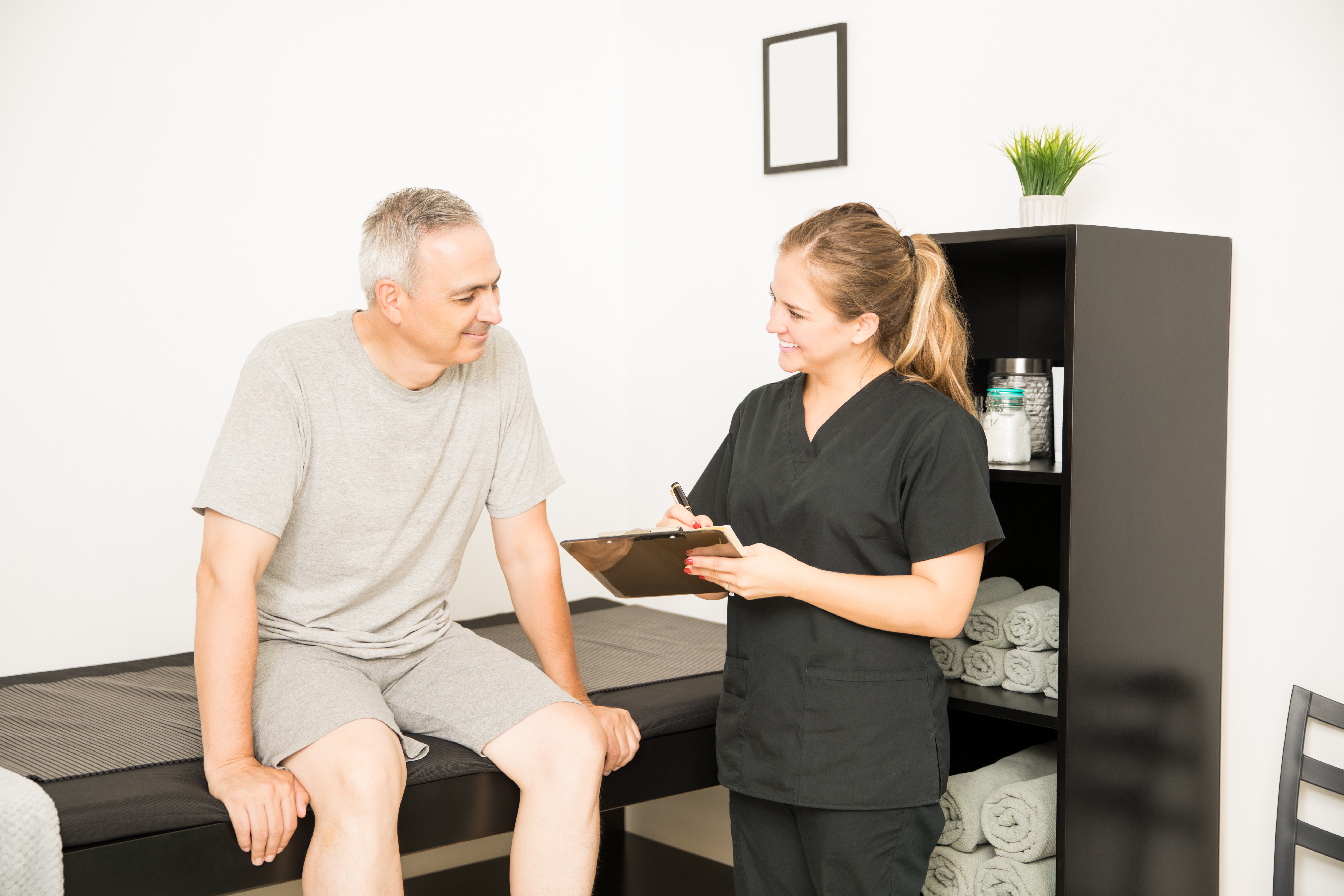 A female healthcare provider in dark scrubs consults with a male patient seated on an exam table in a clinical treatment room.