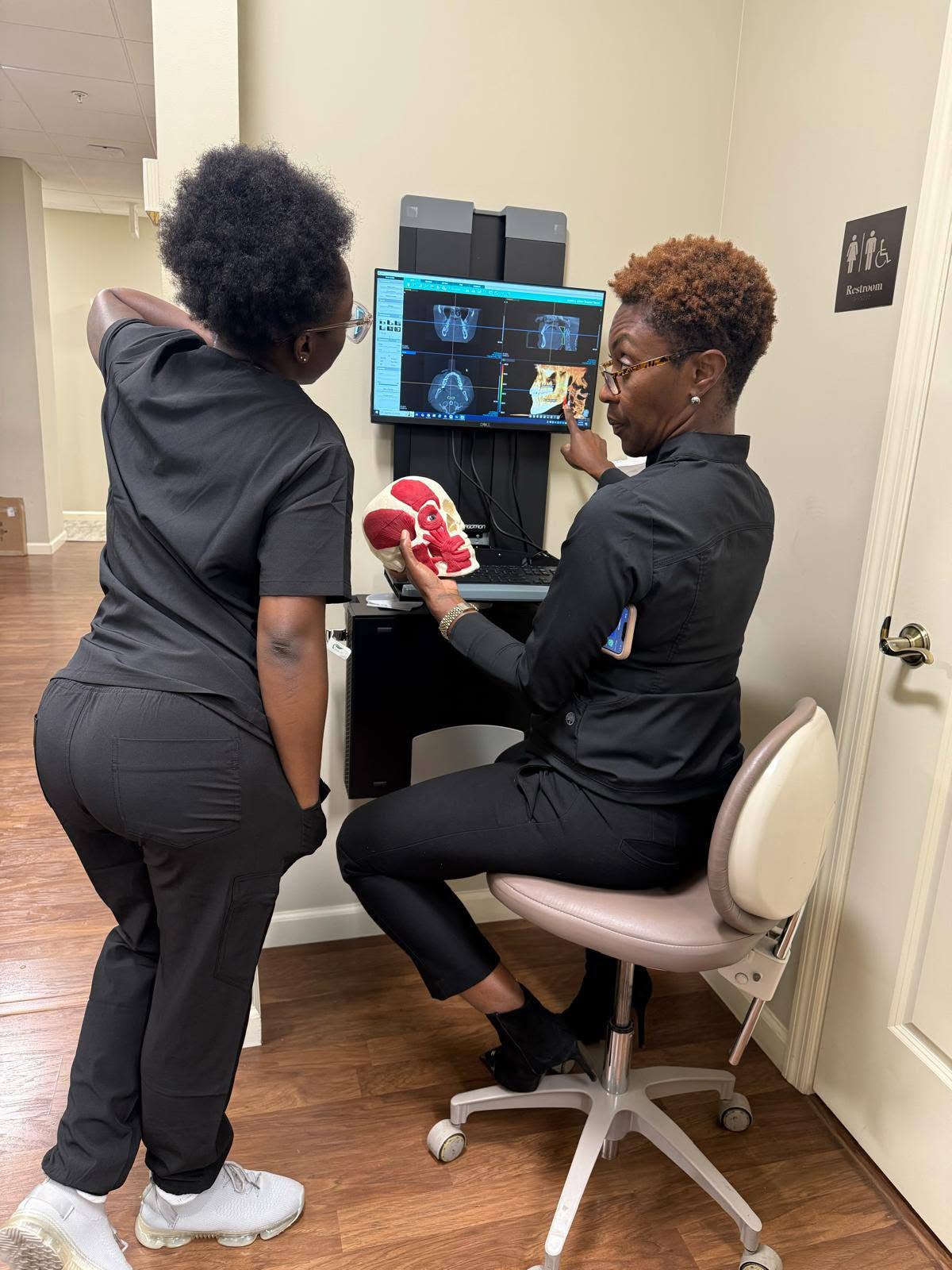 Two team members in black scrubs review a 3D dental CT scan on a monitor while holding a skull model