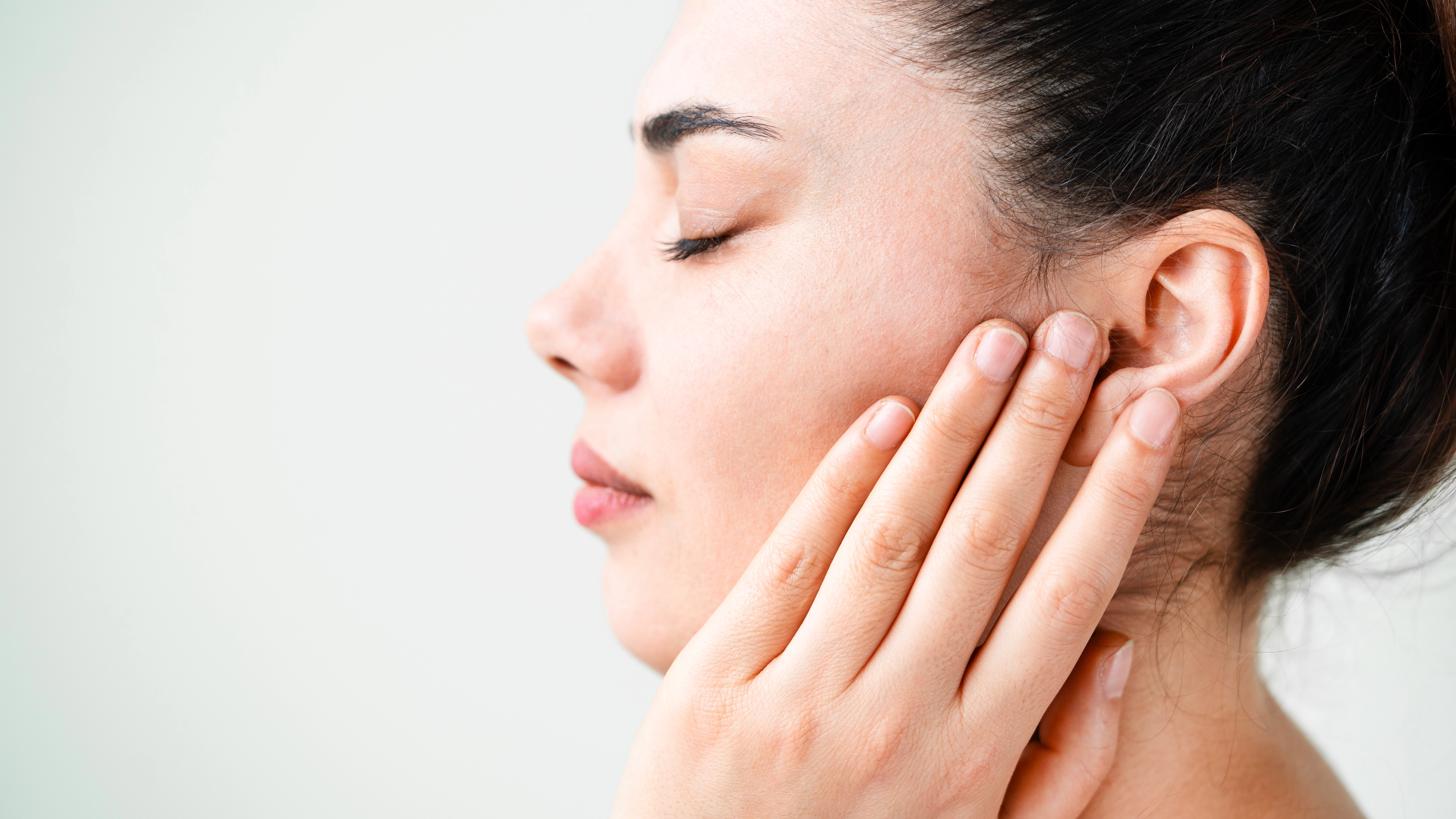 A woman in profile touches her ear and cheek with her eyes closed, highlighting facial and ear skin