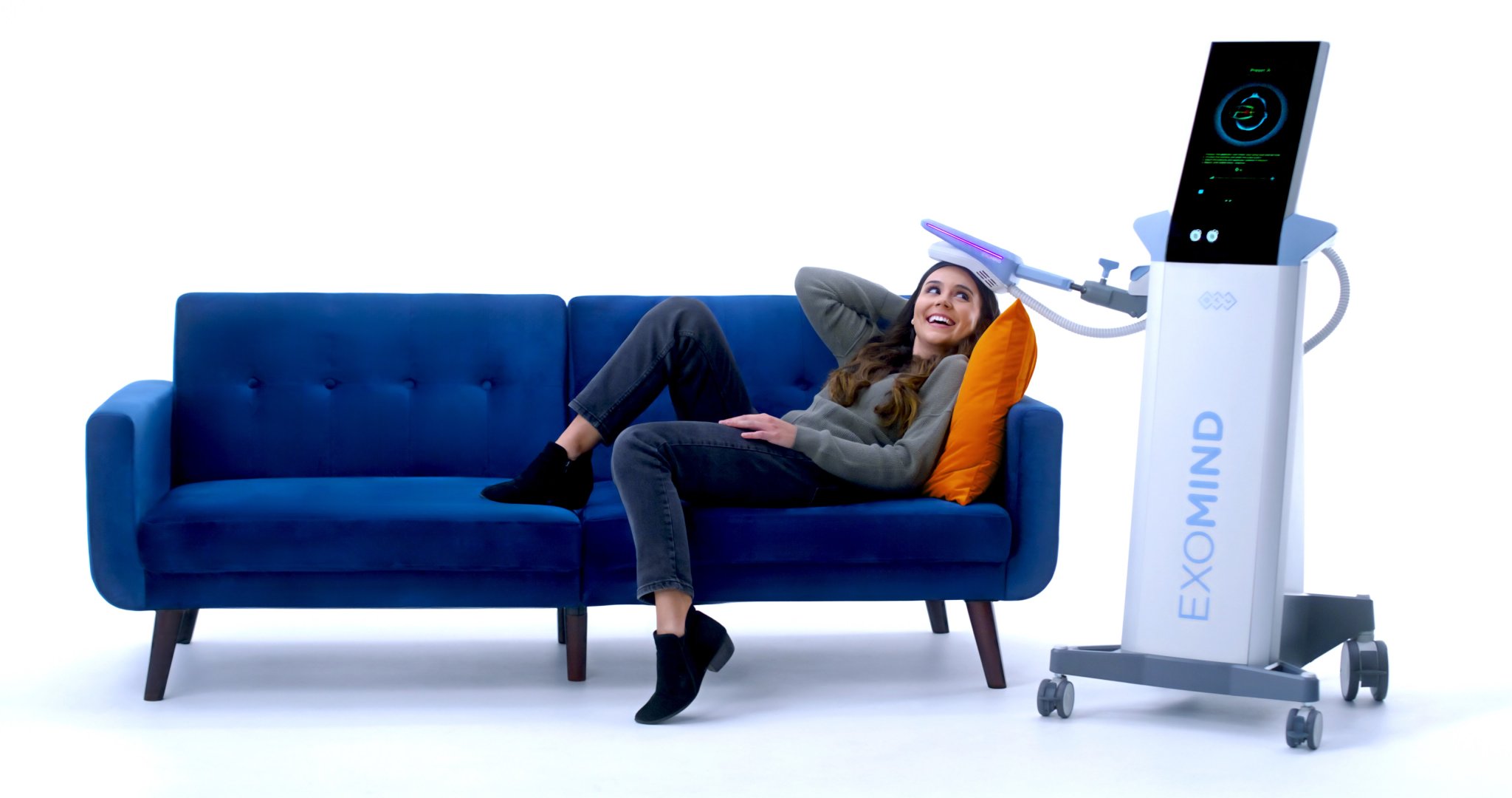 A smiling woman reclines on a blue sofa undergoing a light-based therapy session with a device labeled 'Exomind'