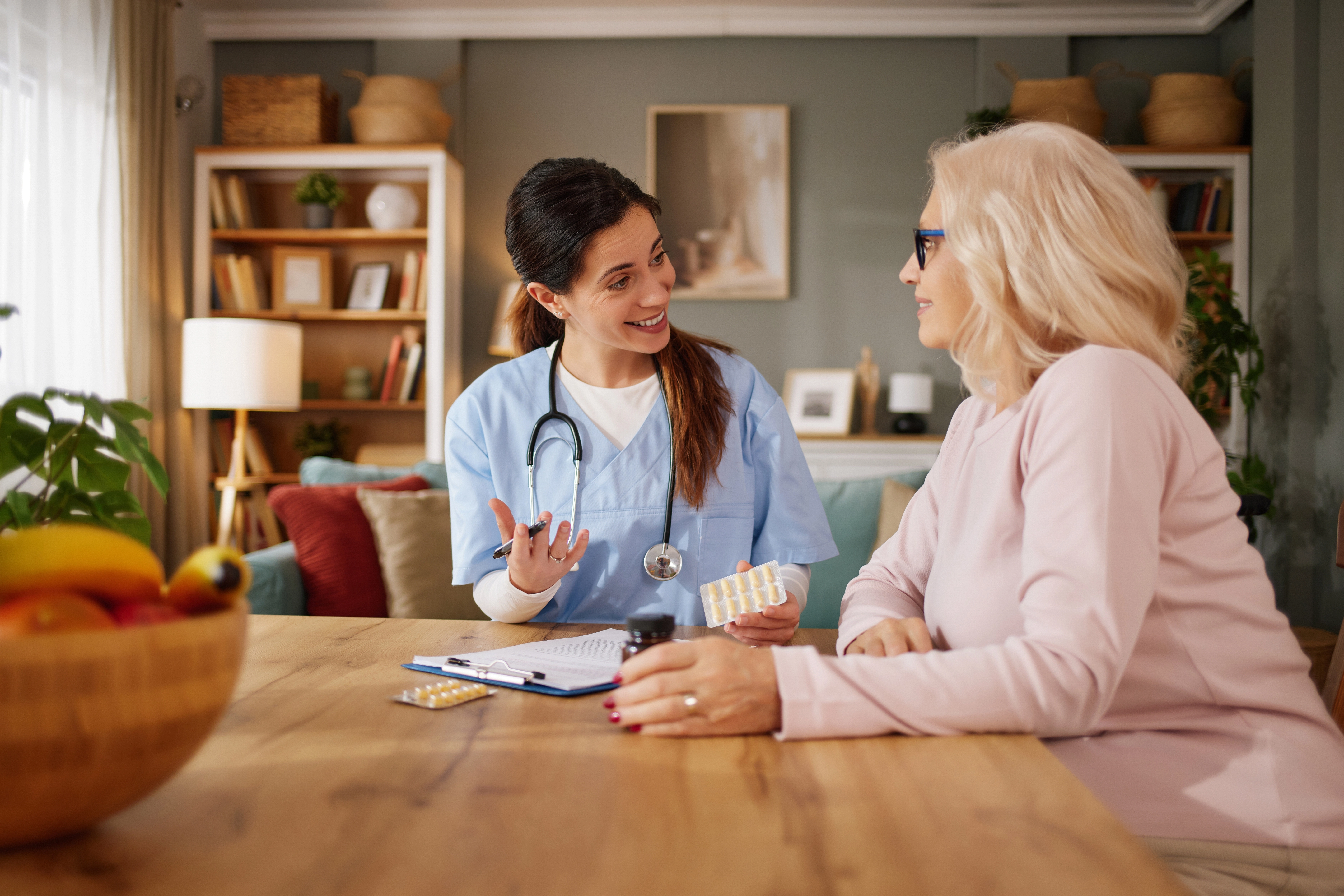 A home health provider in scrubs discusses medication options with a seated older woman during an in-home consultation.