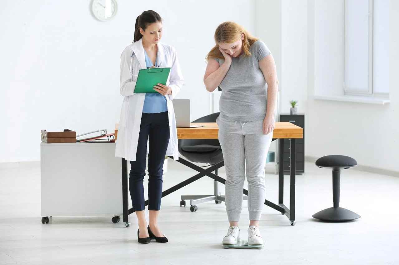 A doctor in a white coat reviews a clipboard while a patient stands on a scale during a weight loss consultation