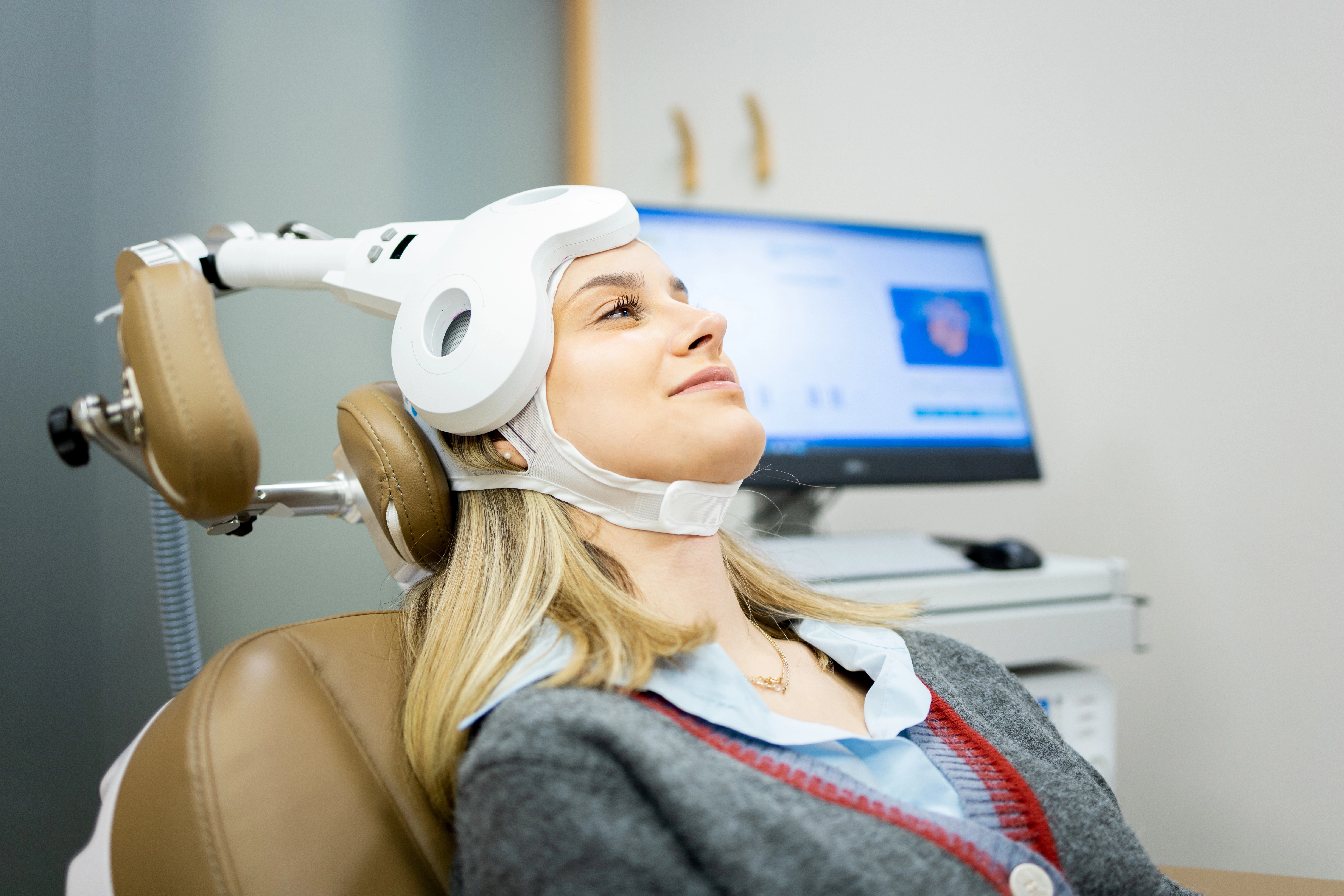 A woman reclines in a treatment chair while undergoing TMS therapy with a magnetic coil positioned against her head