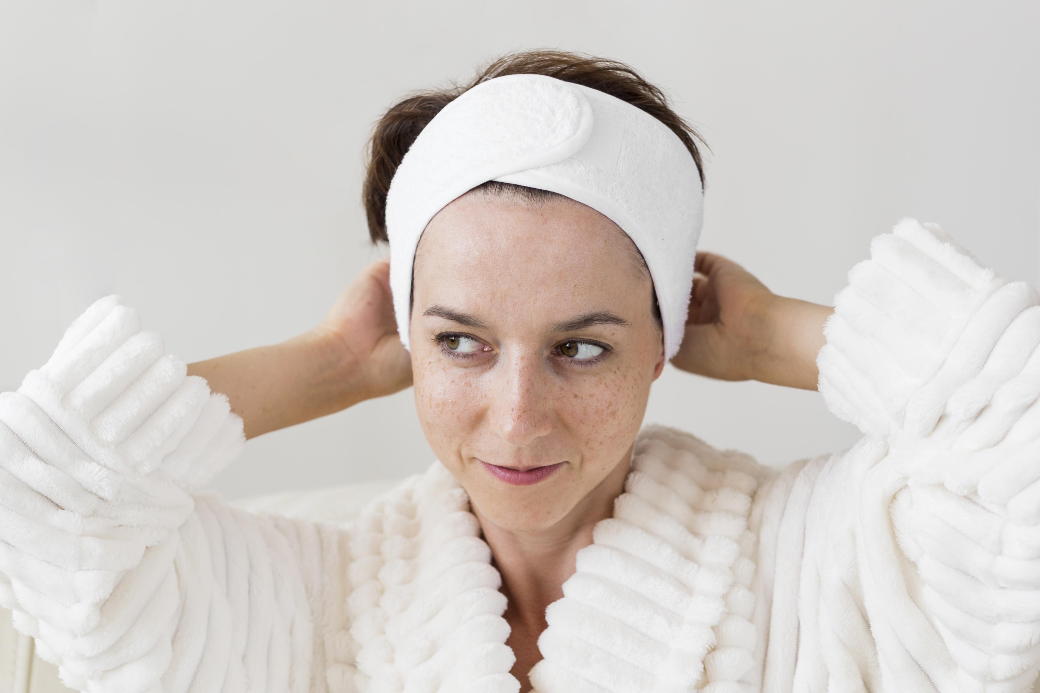 A woman in a white spa robe adjusts her headband while preparing for a skincare treatment
