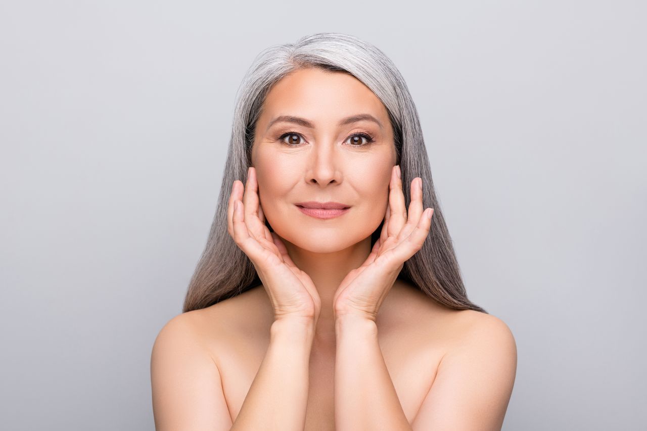 A mature woman with grey hair gently touches her face with both hands, showcasing smooth, healthy-looking skin against a plai