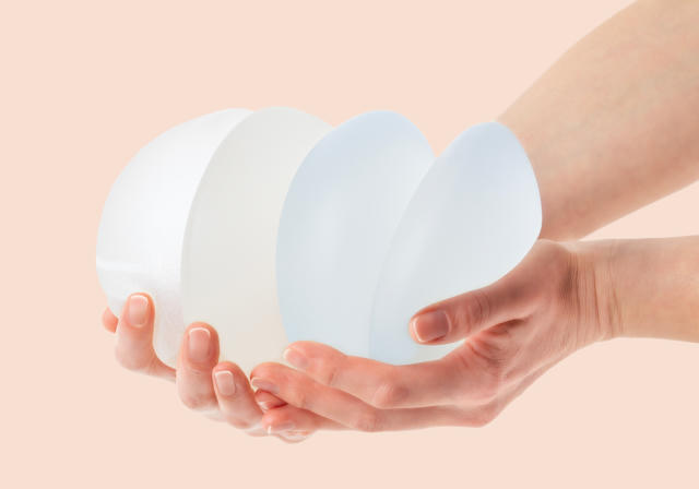 A pair of hands holds various breast implant sizes and shapes against a soft peach background