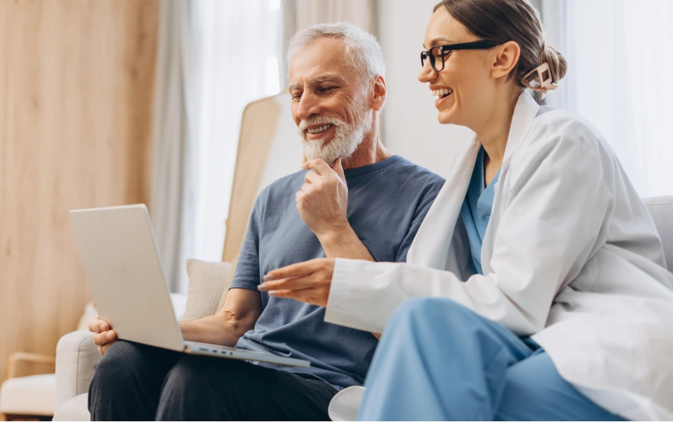 A doctor sits with an older patient on a couch, reviewing information together on a laptop
