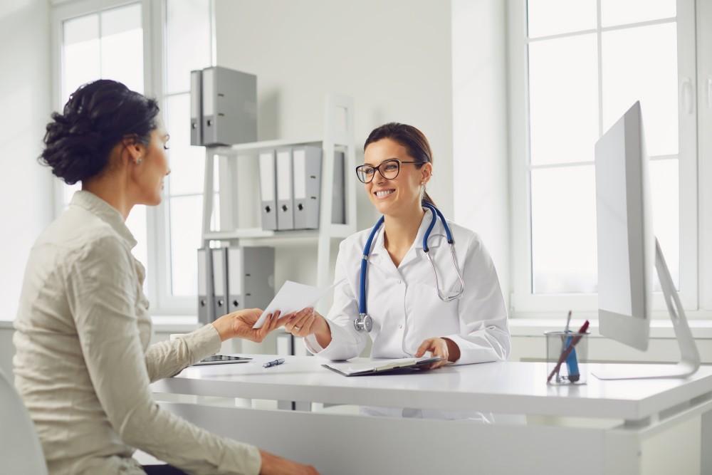 A smiling female doctor in a white coat hands a document to a patient during a consultation in a bright office.