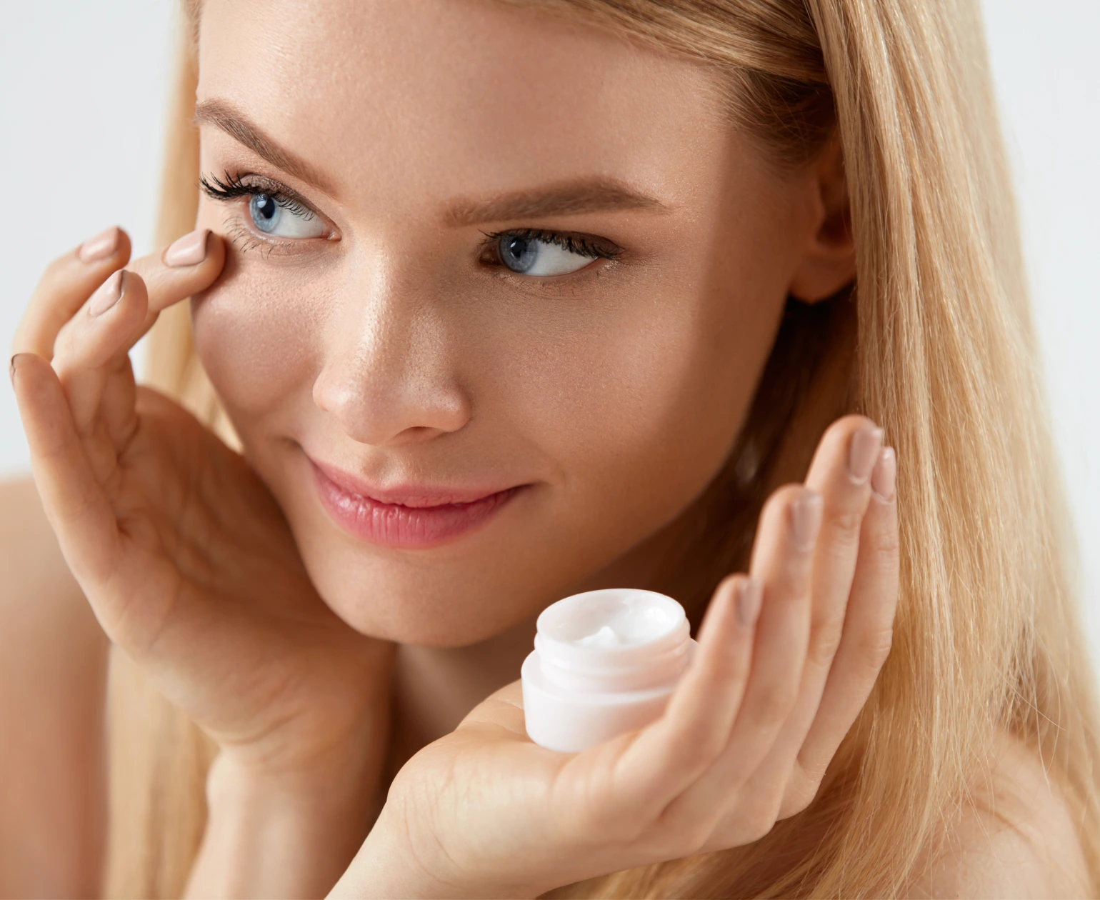 A young woman with blonde hair applies eye cream from a small jar while touching under her eye