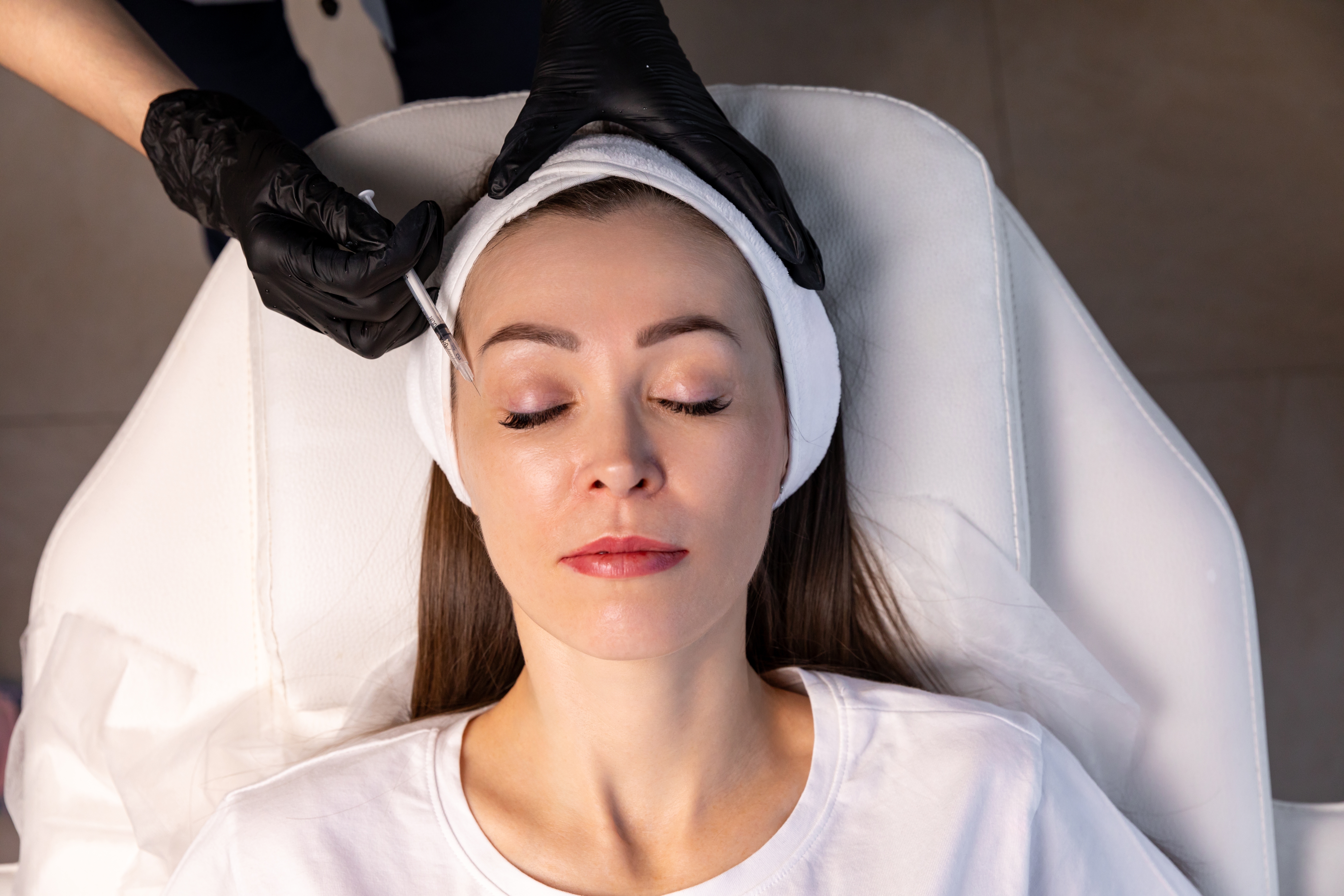 A woman reclines with eyes closed as a gloved provider administers a facial injection, likely Botox, in a medspa treatment ro