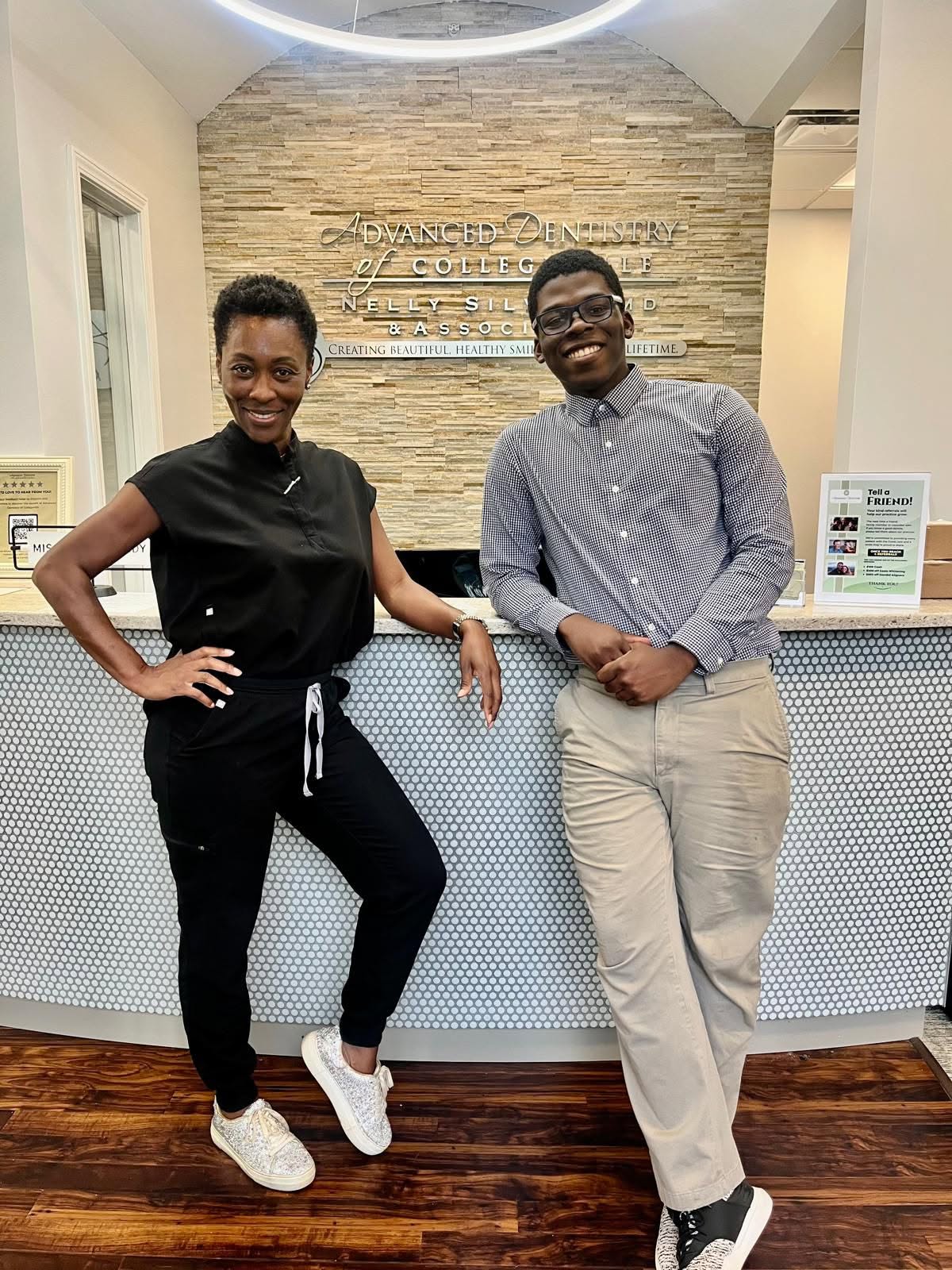 Two members of our dental team smiling at the front desk of our office, with our sign on the stone wall behind them