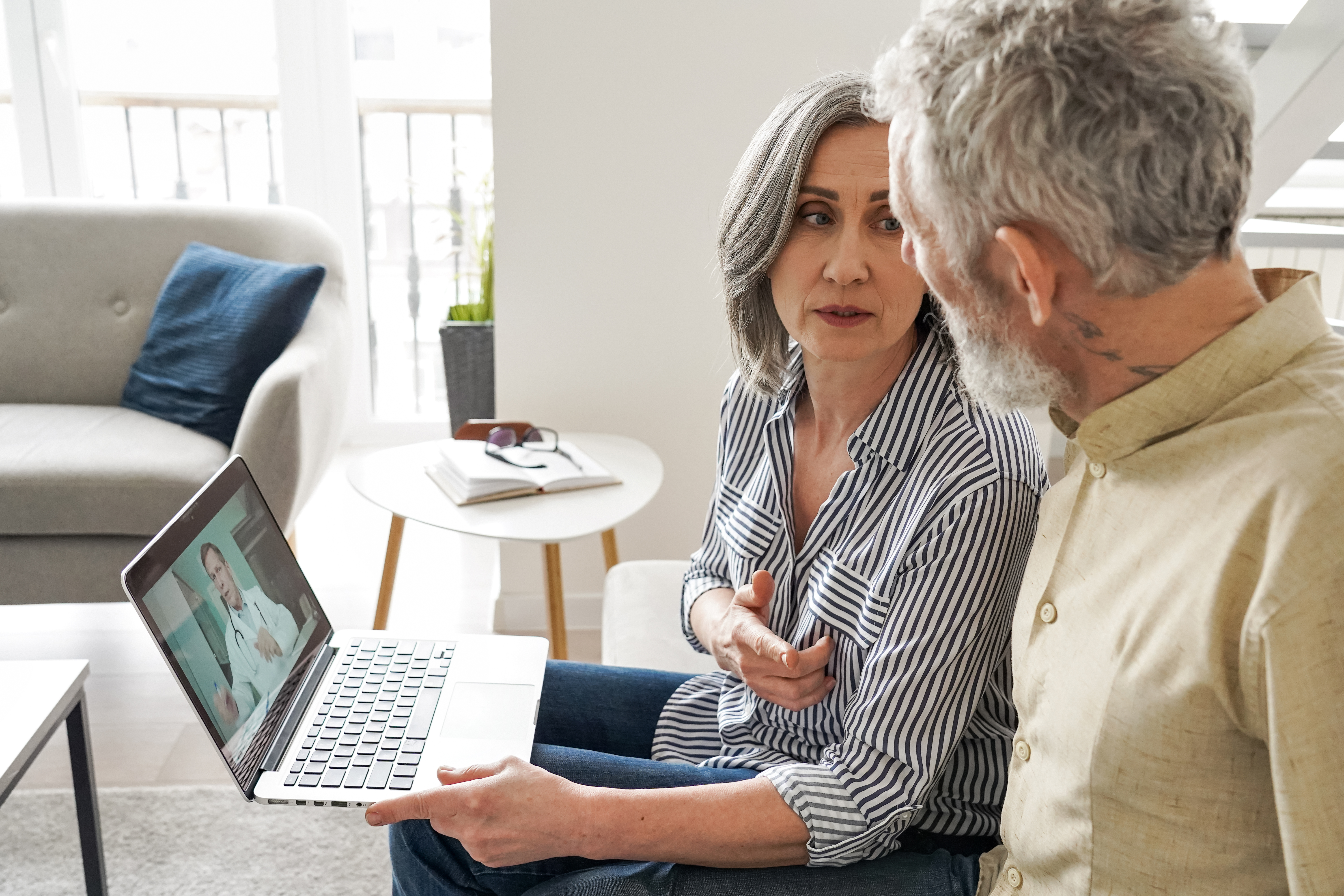 An older couple sits together on a sofa engaging in a telehealth video call with a physician on a laptop