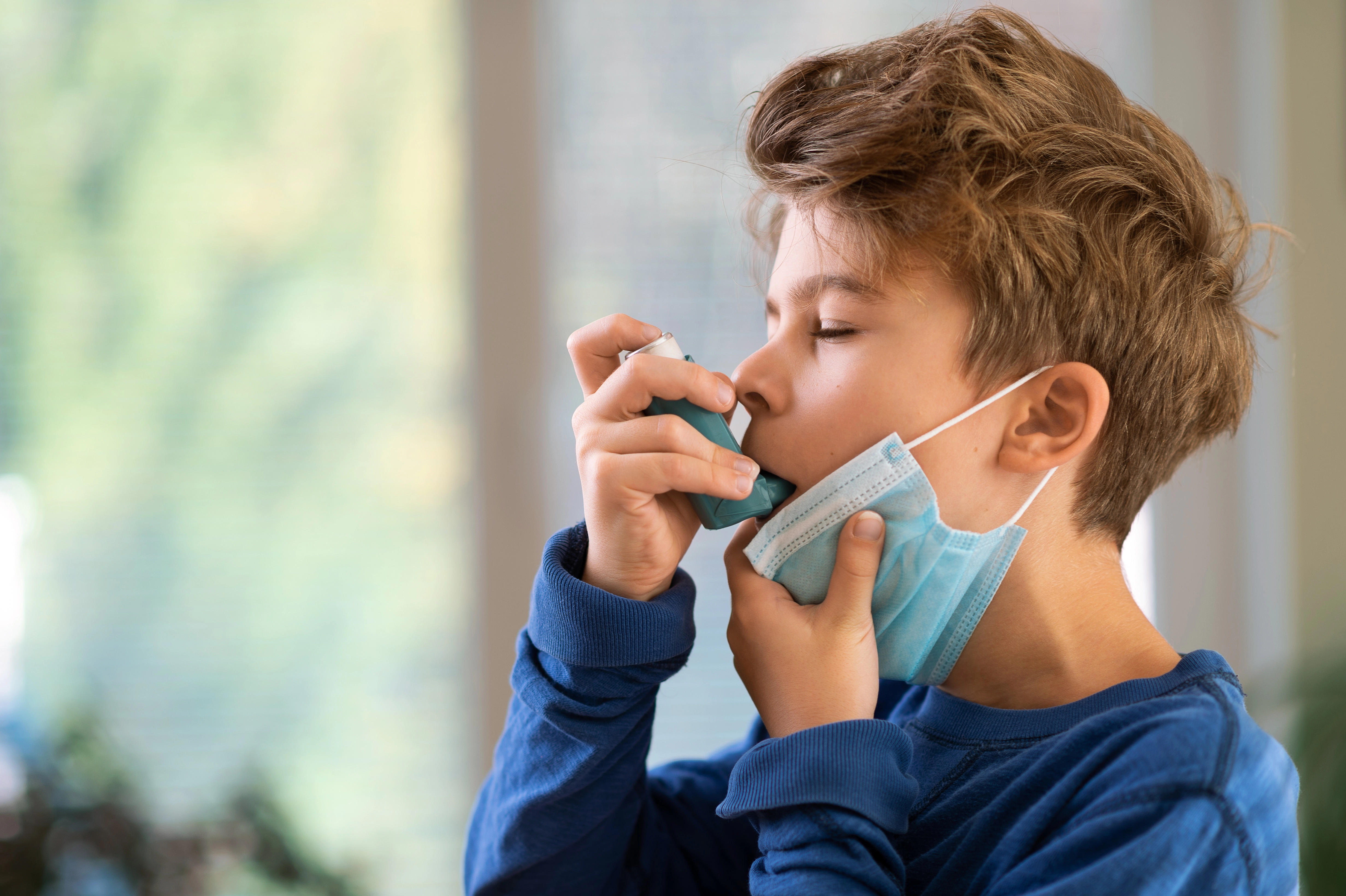 A young boy pulls down his face mask to use an asthma inhaler