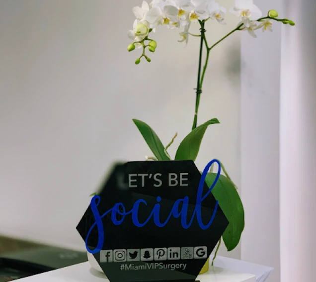 A Miami VIP Surgery social media sign beside a white orchid in our South Miami office