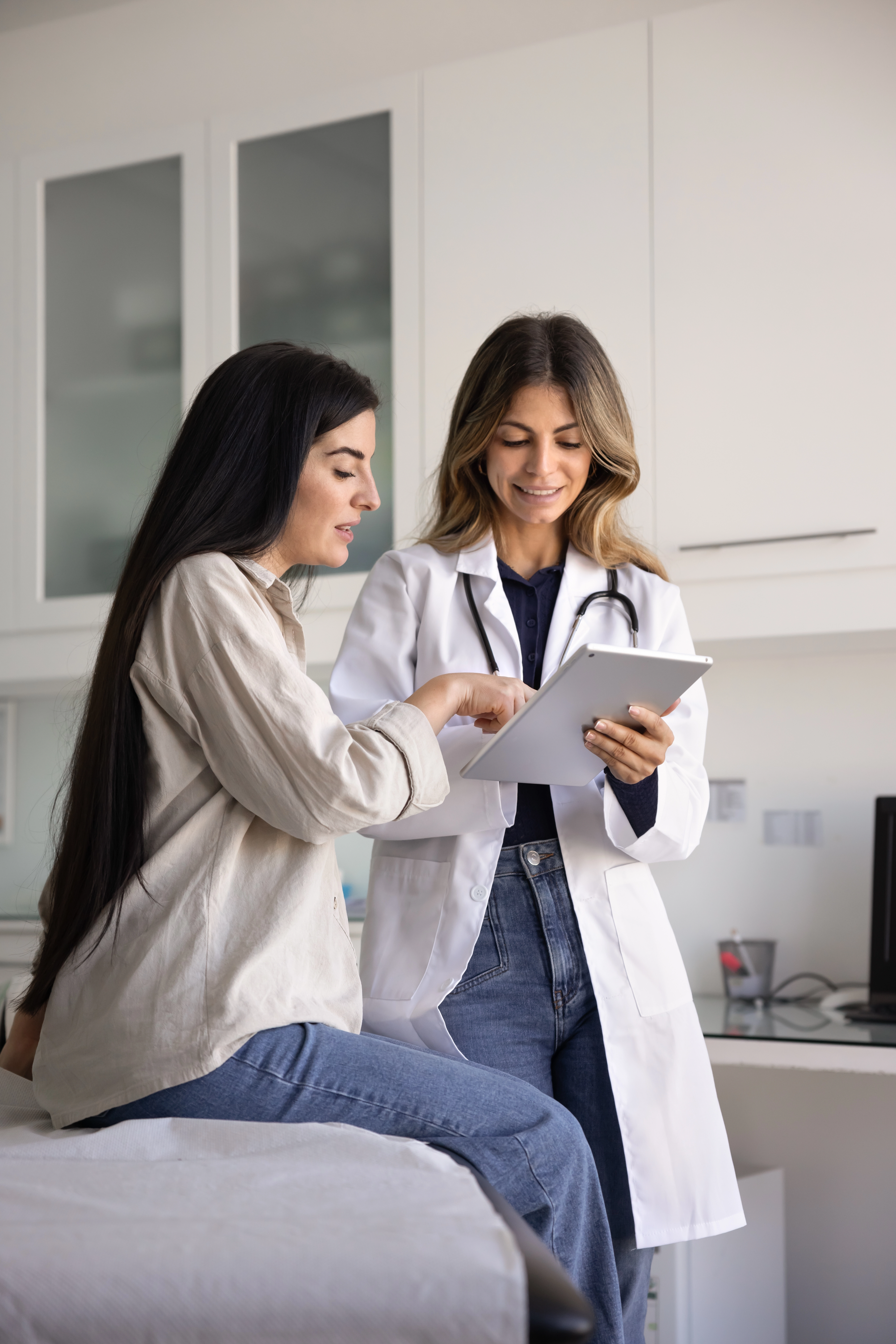 A doctor in a white coat reviews information on a tablet with a seated patient in a clinical exam room, illustrating a discus