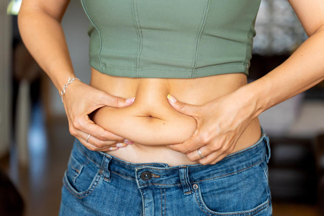 A woman pinches excess fat on her stomach while wearing jeans and a cropped top