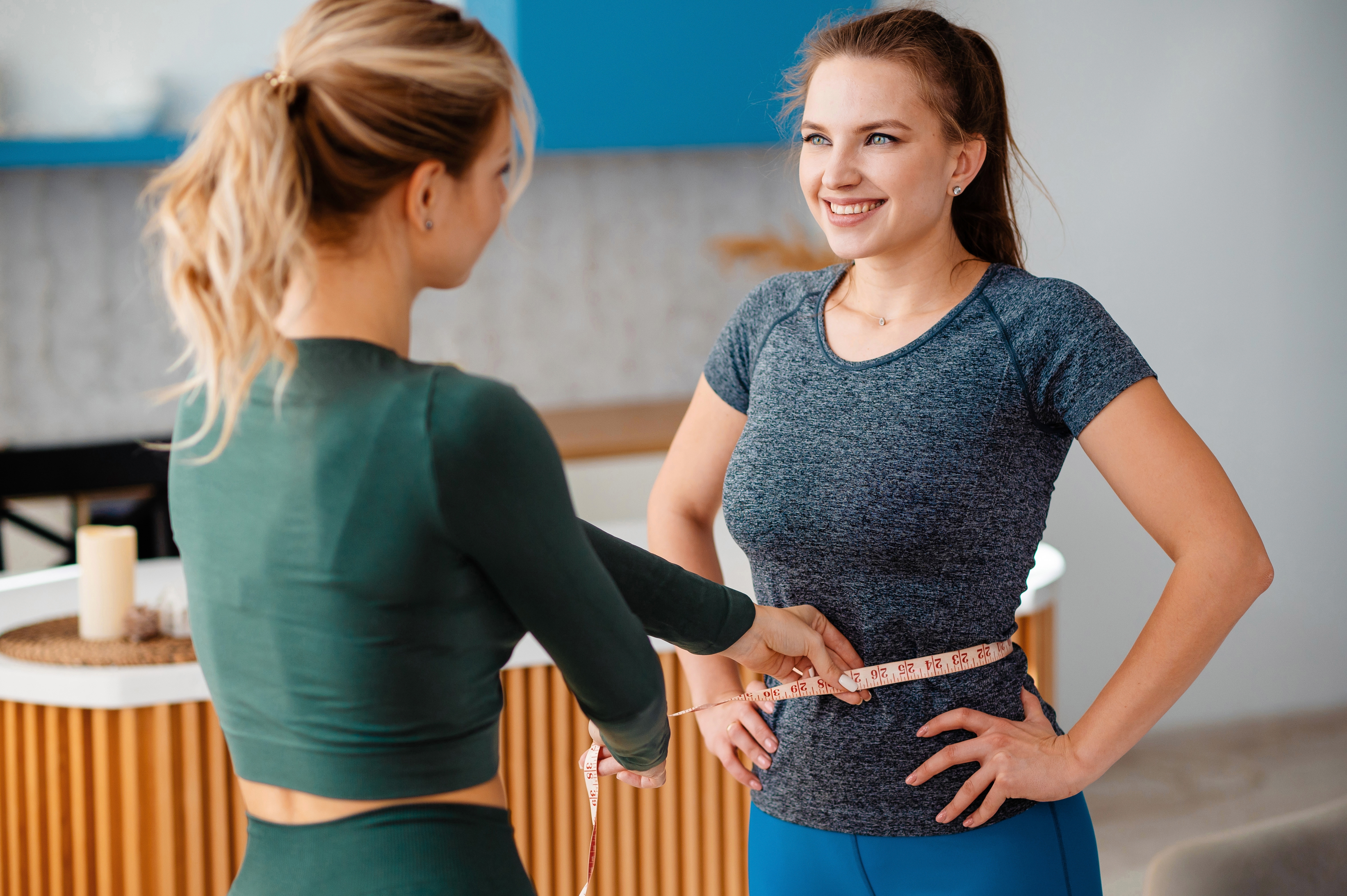 A health coach measures a smiling woman's waist with a tape measure to track weight loss progress