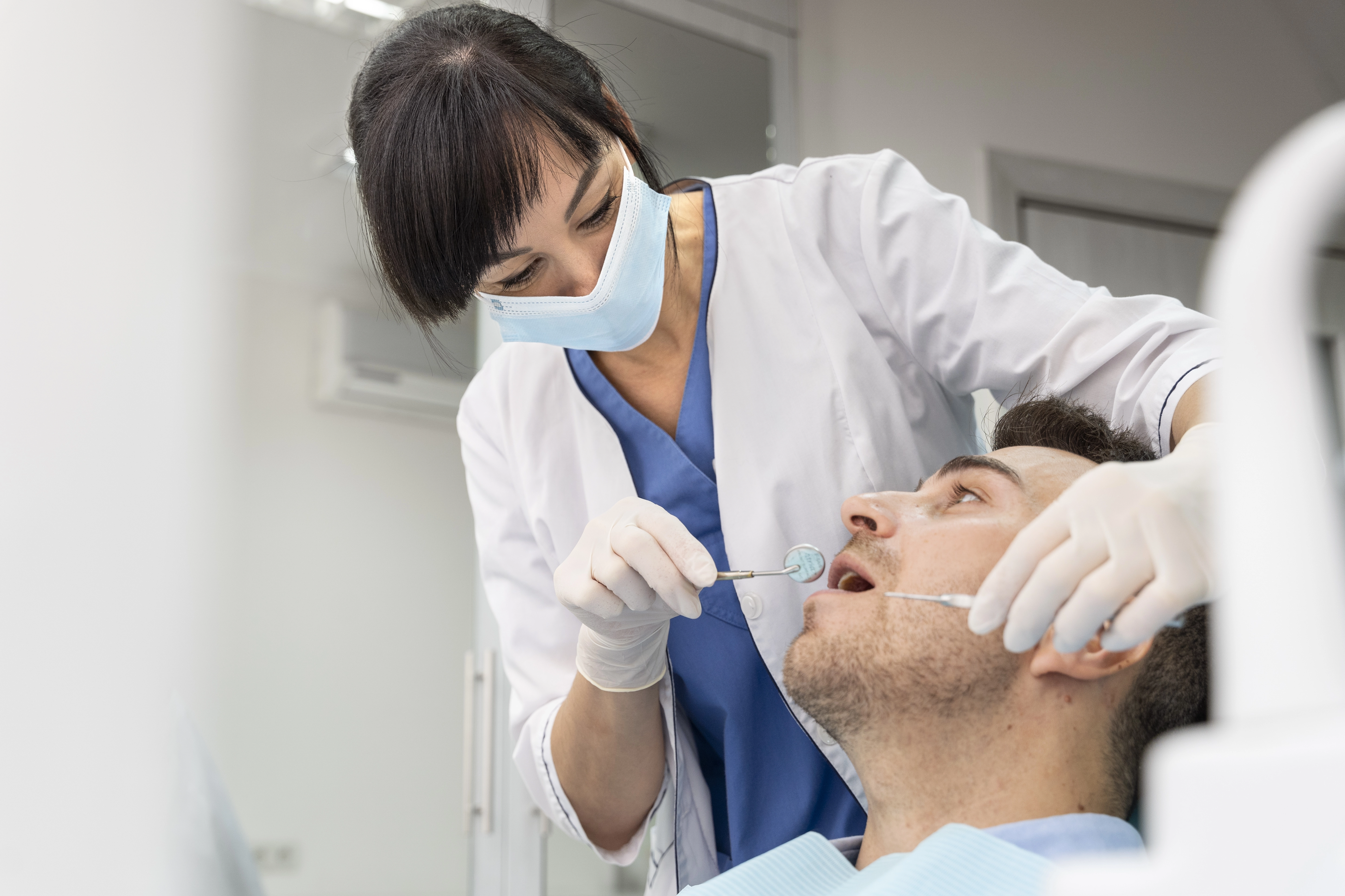 A female dentist wearing a mask and gloves examines a male patient's teeth using a dental mirror