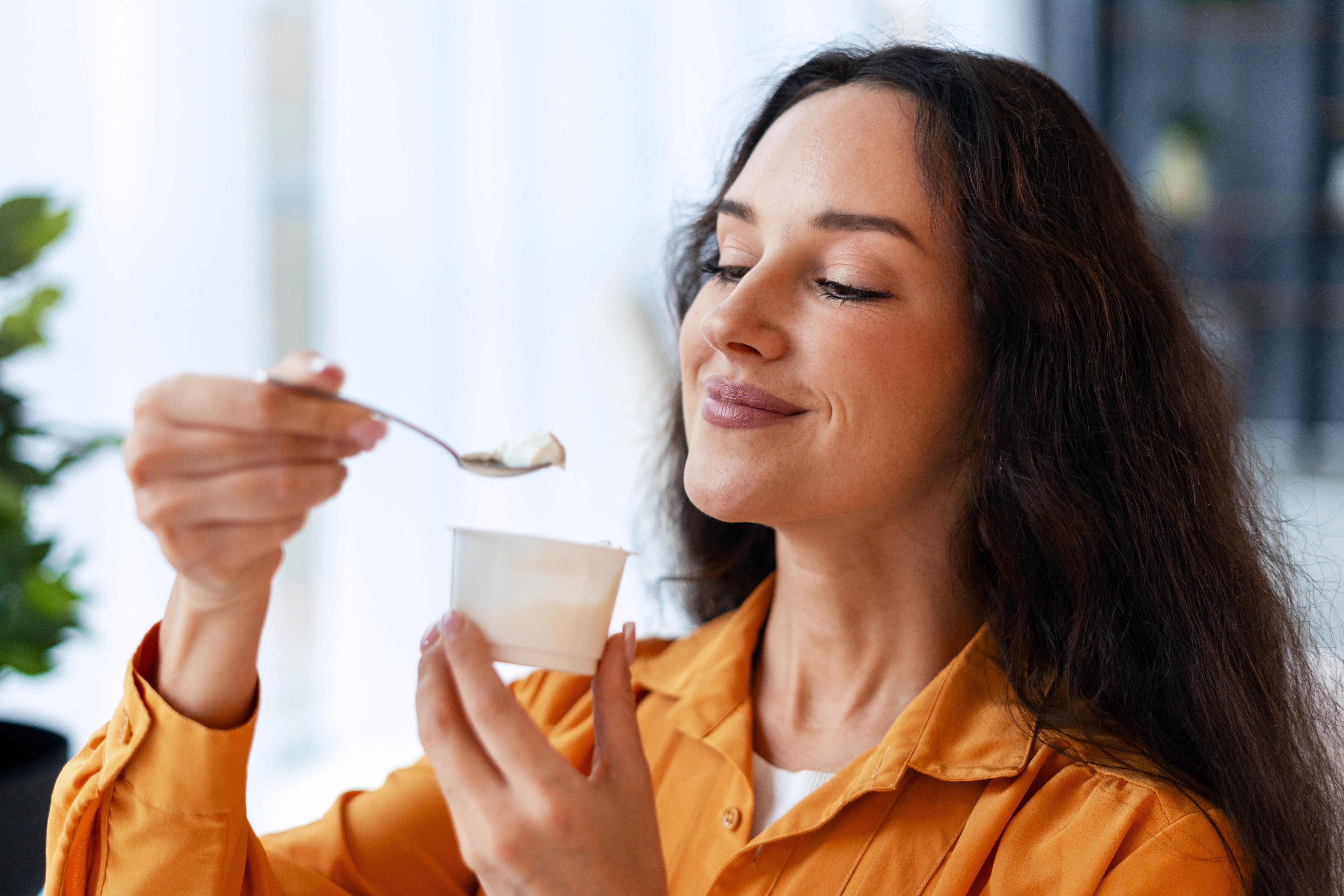 Woman in an orange shirt enjoying a spoonful of yogurt in a bright room