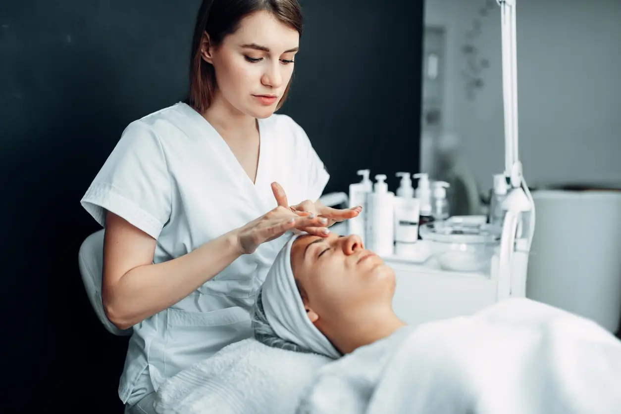 A esthetician in white scrubs performs a facial skin treatment on a relaxed client wrapped in a towel, surrounded by skincare
