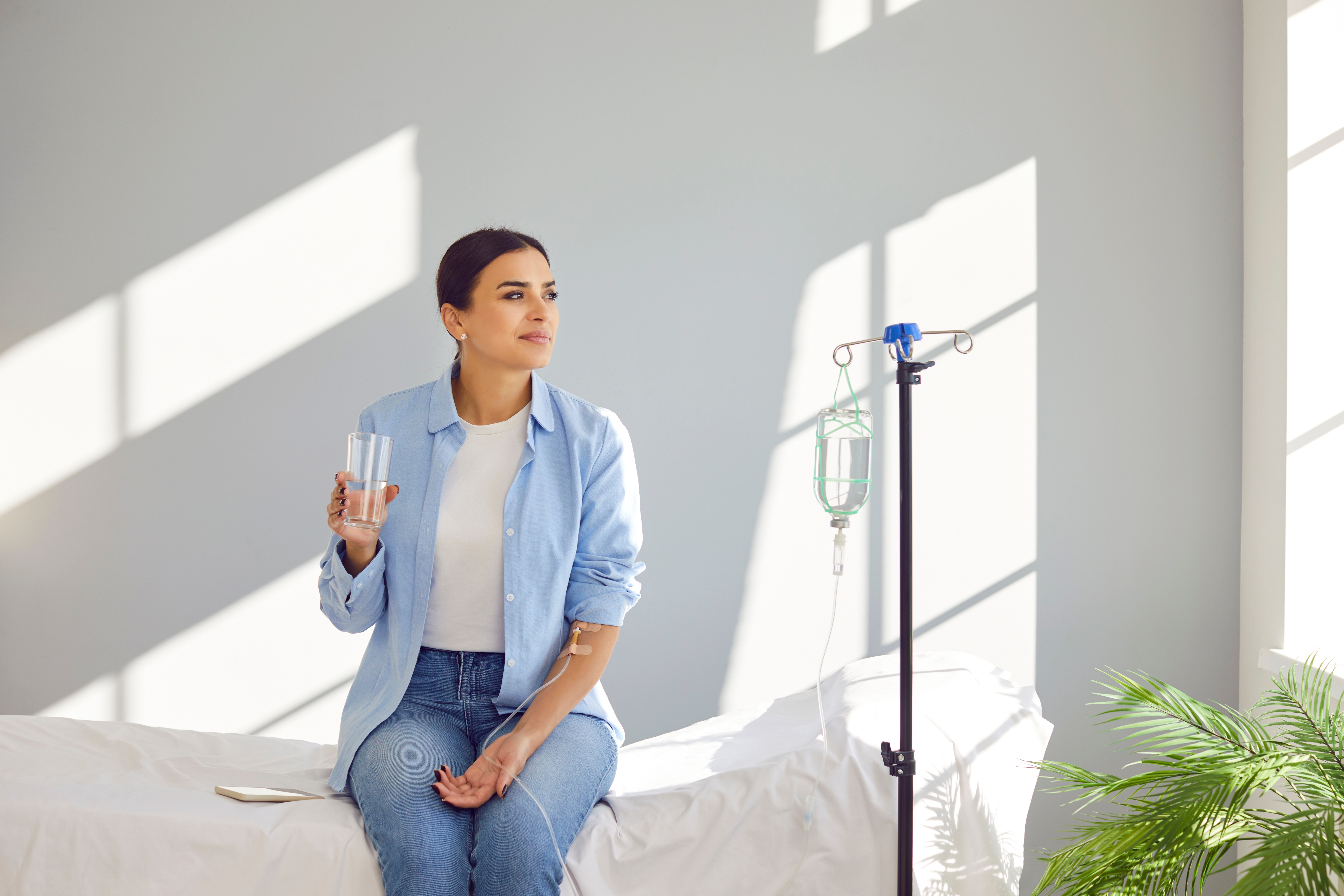 Woman sitting on a treatment bed with a glass of water while an IV drip runs into her arm in a sunlit room