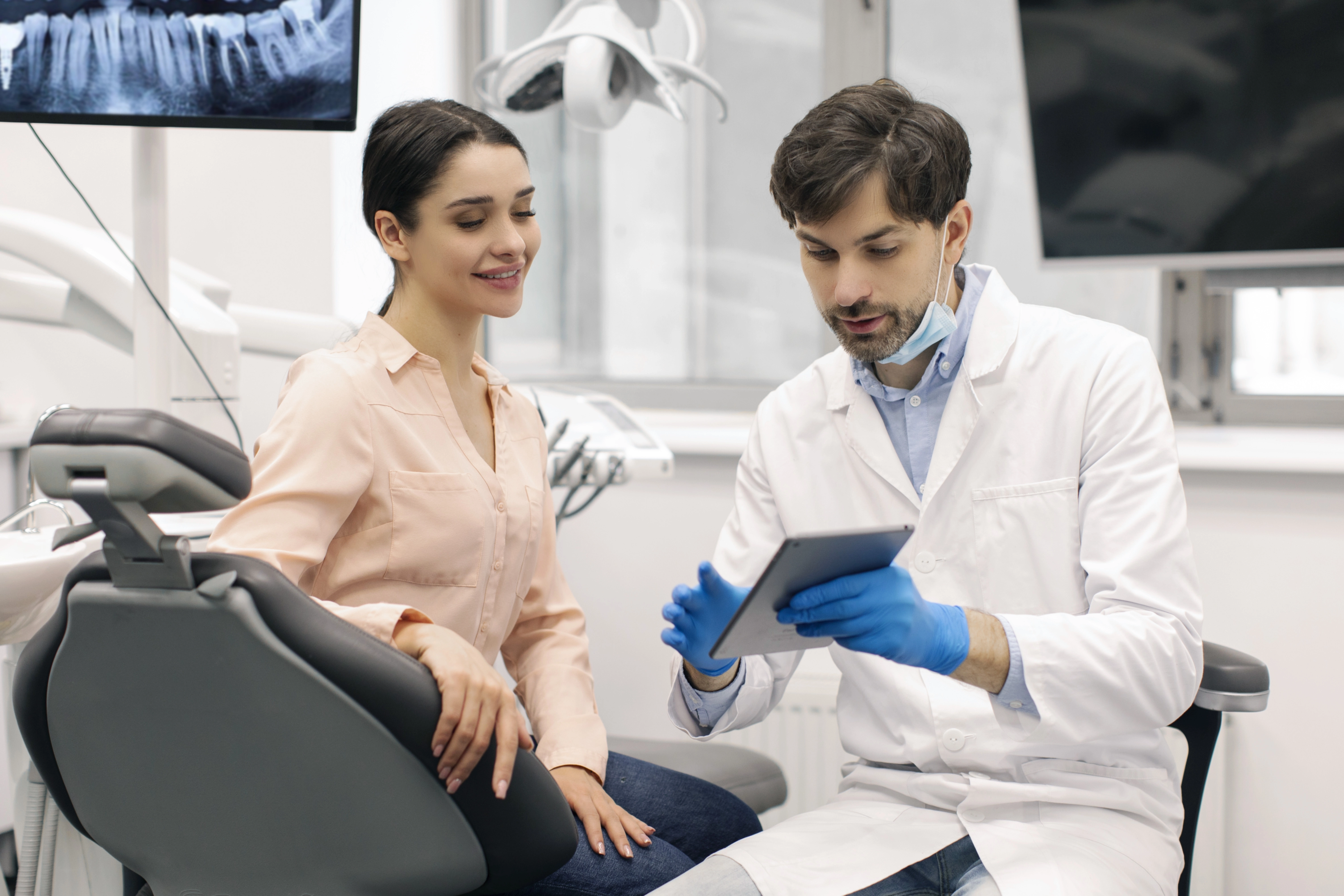 A male dentist in a white coat and blue gloves reviews information on a tablet with a smiling female patient seated in a dent