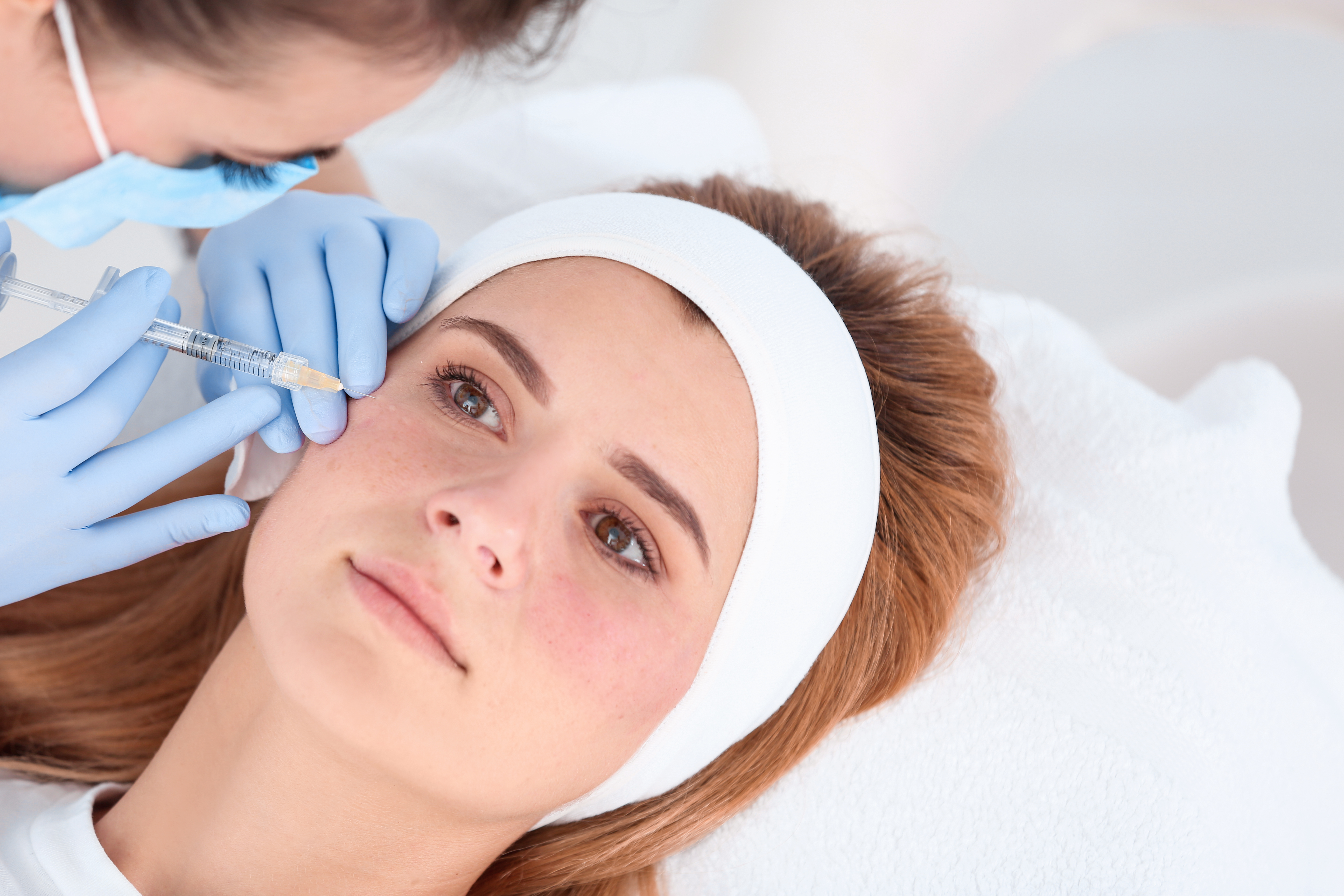 A gloved provider administers a dermal filler injection near a woman's under-eye area during a medspa treatment