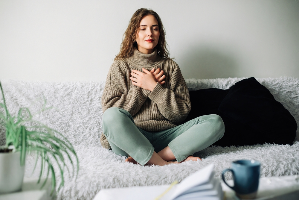 Woman sitting cross-legged with eyes closed and both hands resting over her heart in a calm, bright room