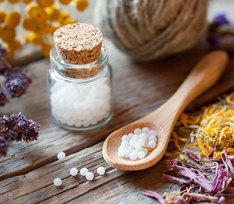 Glass jar and wooden spoon of homeopathic globules among dried herbs and flowers