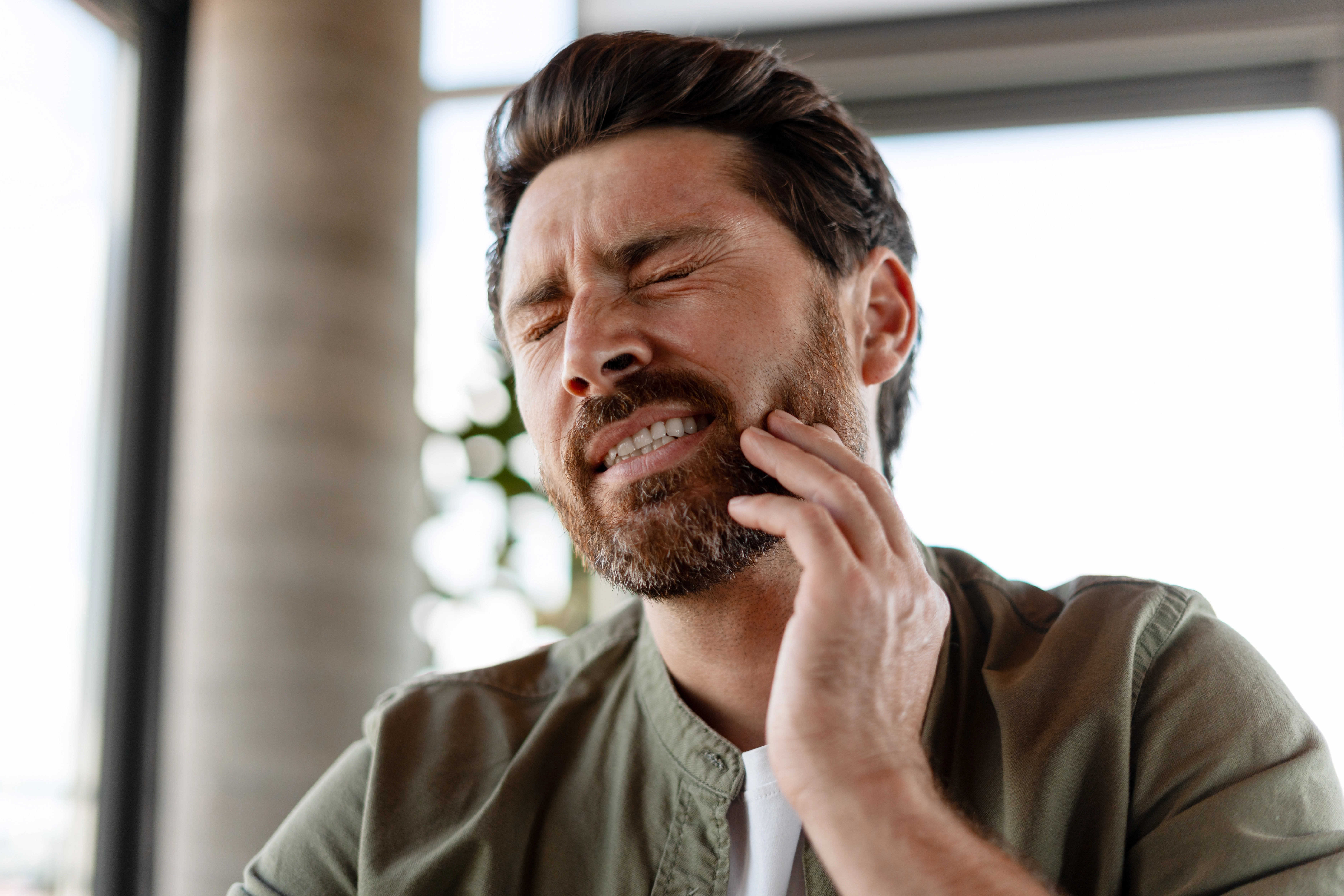 A man winces in pain while touching his jaw, indicating tooth or dental discomfort