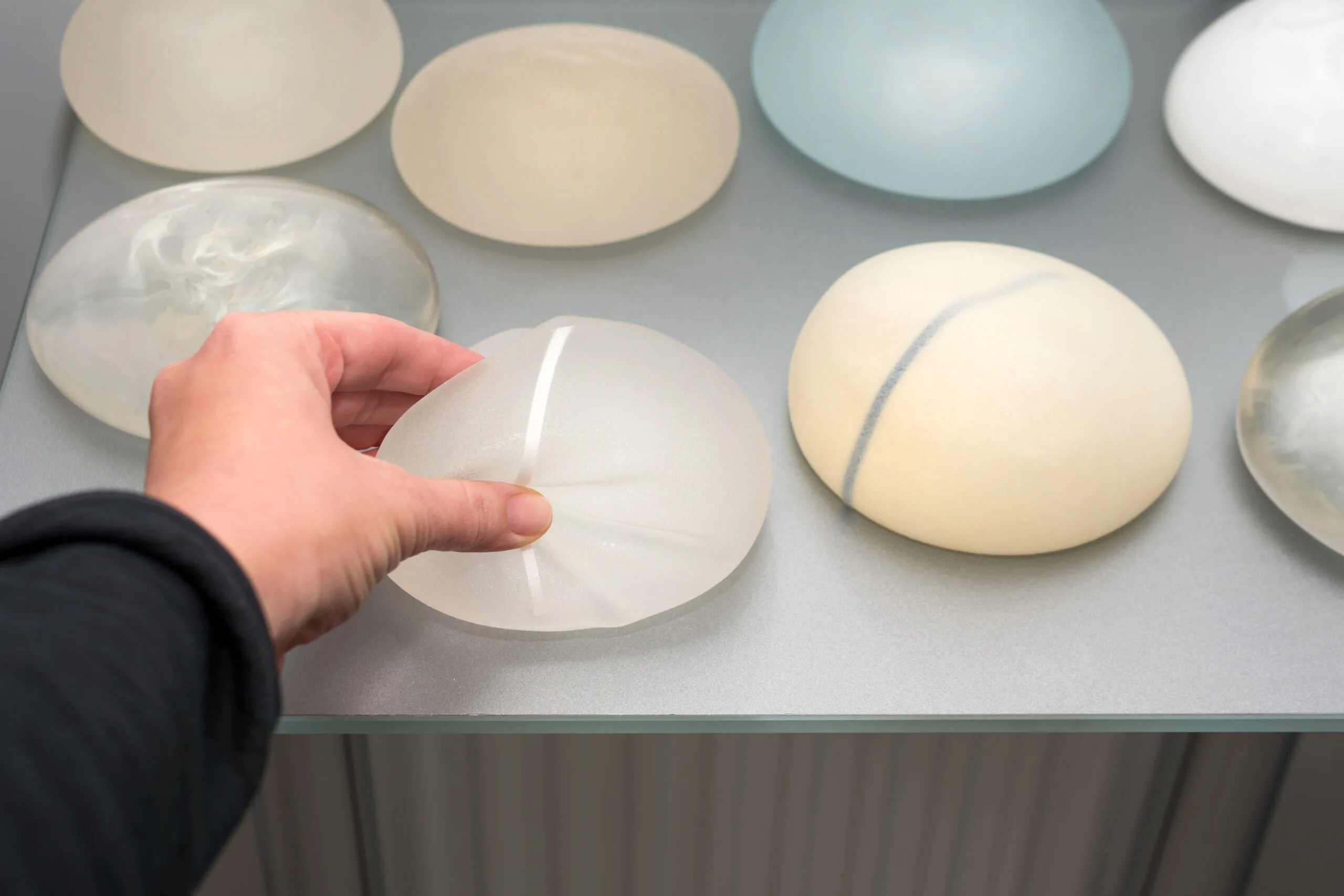 A hand holds up a silicone breast implant sample next to a row of other implant options displayed on a table