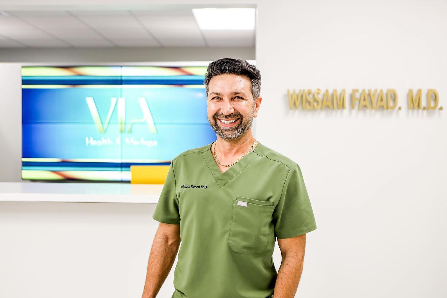 Dr. Wissam Fayad in green scrubs smiling in front of the VIA Health & Medspa logo wall