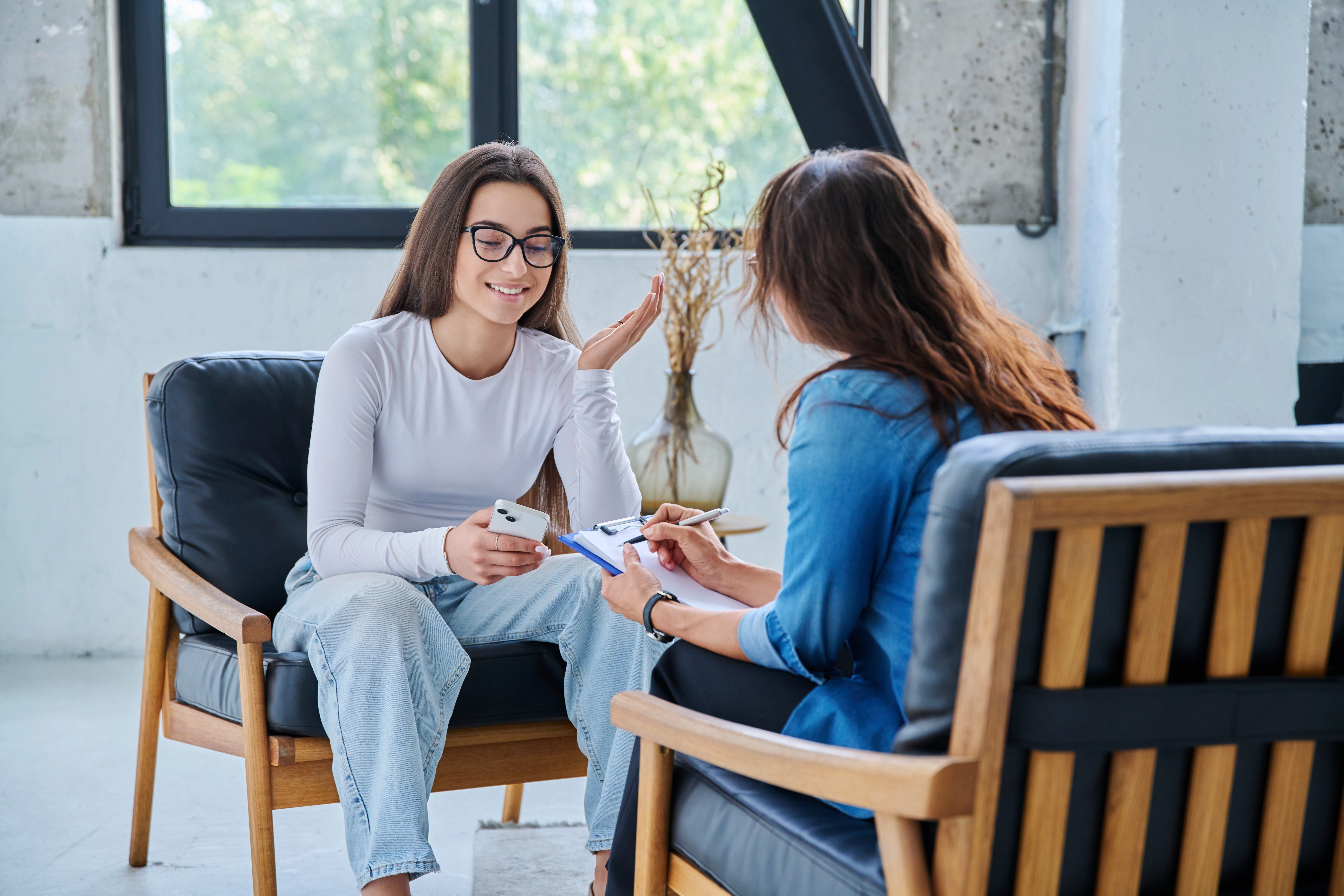 A young woman smiles while talking with a therapist who takes notes on a clipboard during a counseling session