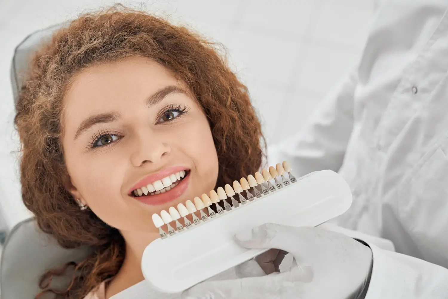 A smiling female patient in a dental chair has her tooth shade checked against a shade guide by her dentist